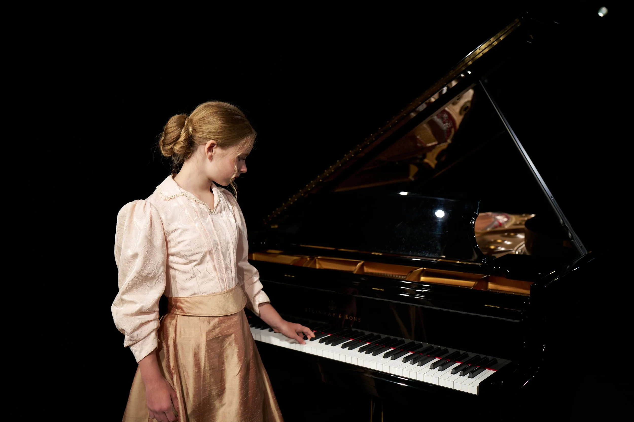 A young girl with red hair styled in a braid, wearing a cream and beige vintage dress, playing a grand piano on a black background.