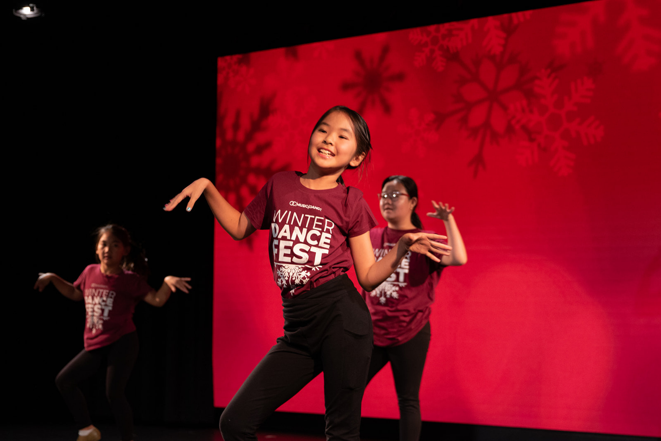 Two women are performing a dance or yoga move on a black stage, with one wearing a red jacket and the other wearing a purple jacket, both in a plank position with their heads turned to the side.