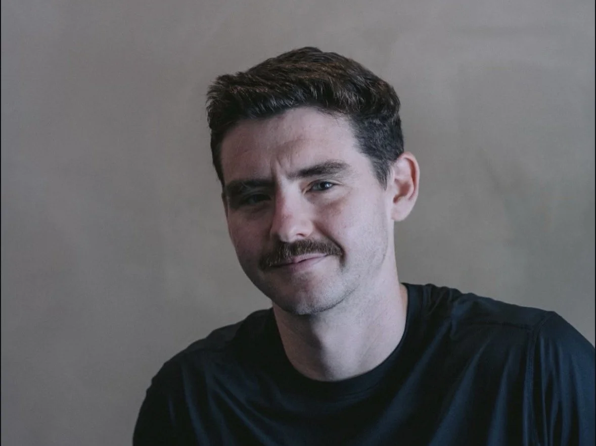 Headshot of a young man with short dark hair and a mustache, wearing a black shirt, against a plain background.