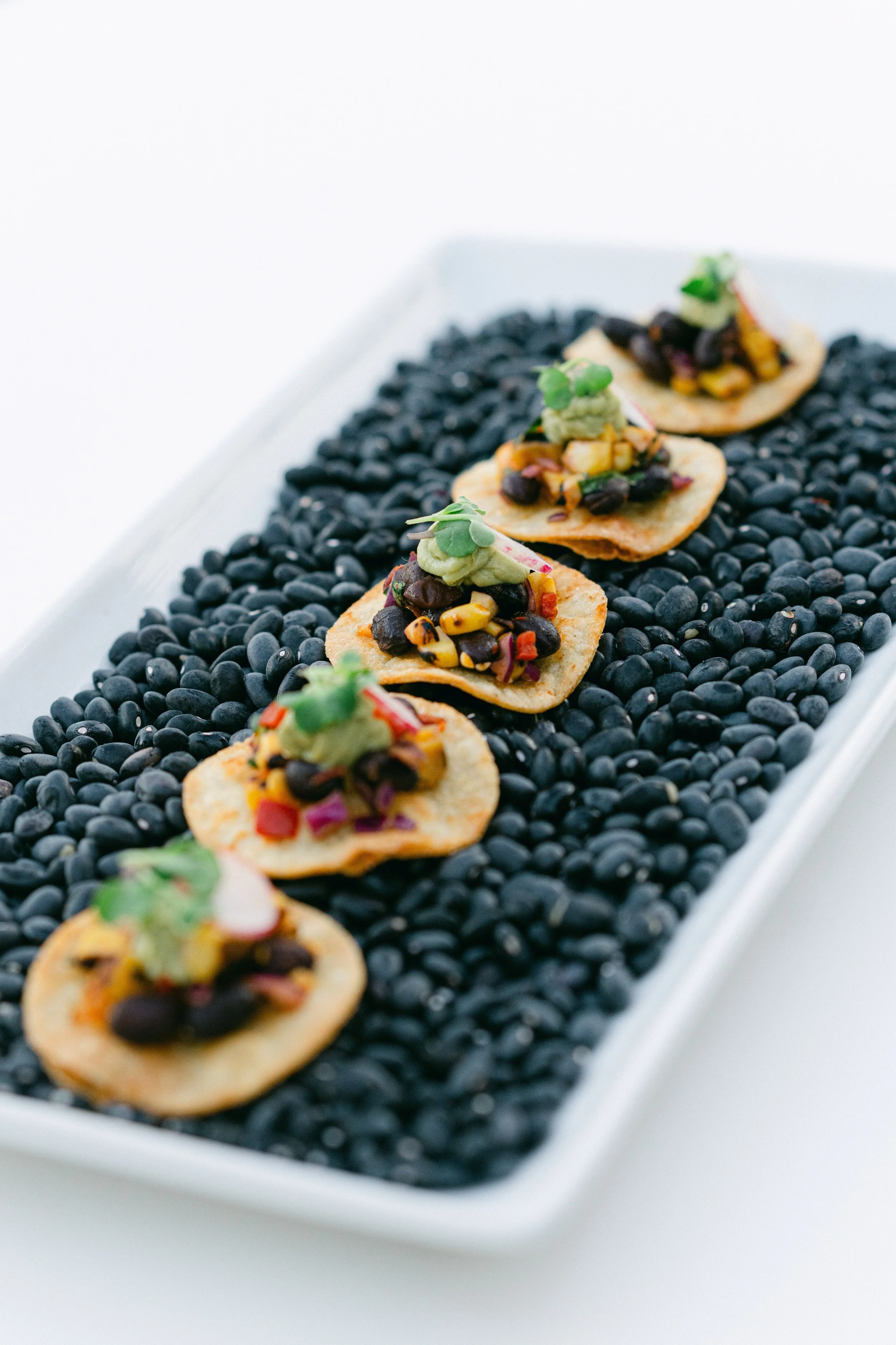 Five tortilla chips topped with black beans, guacamole, and colorful chopped vegetables, arranged in a row on a rectangular black tray filled with black beans.