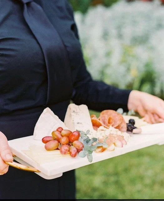 A person holding a cheese and charcuterie board outdoors, featuring three types of cheese, grapes, and cured meats, with greenery in the background.