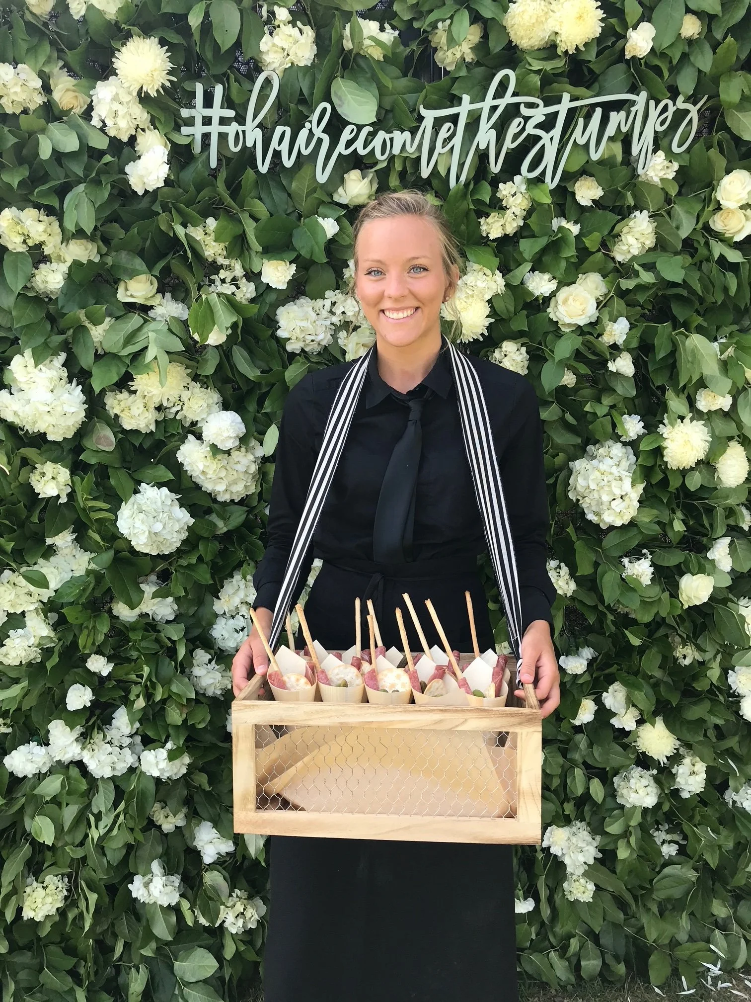 A smiling woman in black attire holding a wooden crate filled with paper cups topped with snacks, standing in front of a green leafy wall decorated with white flowers and a sign that reads '#hairecommendthebestmups'.