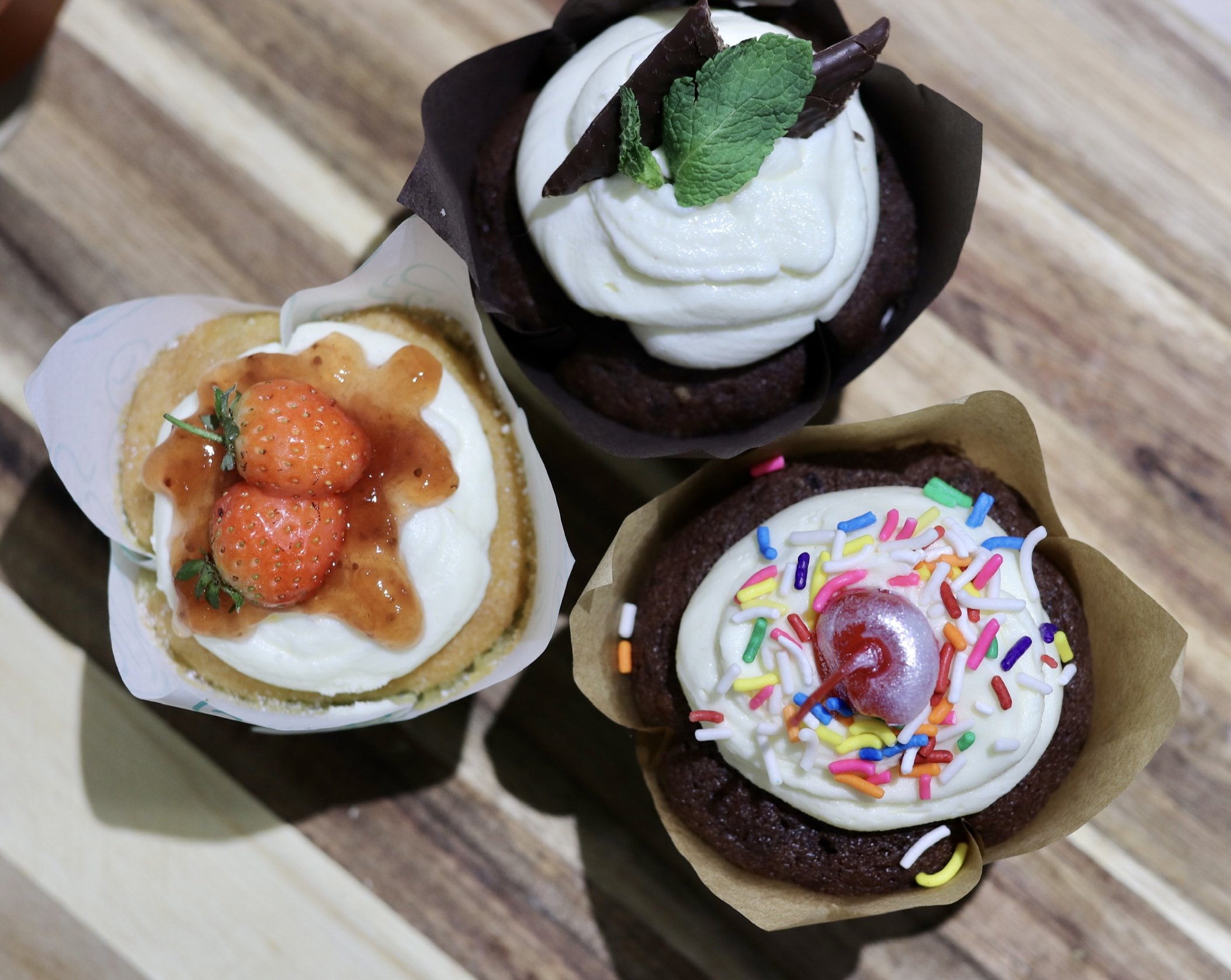 Three cupcakes with different toppings: strawberry with sauce, chocolate with mint, and sprinkles with a cherry.