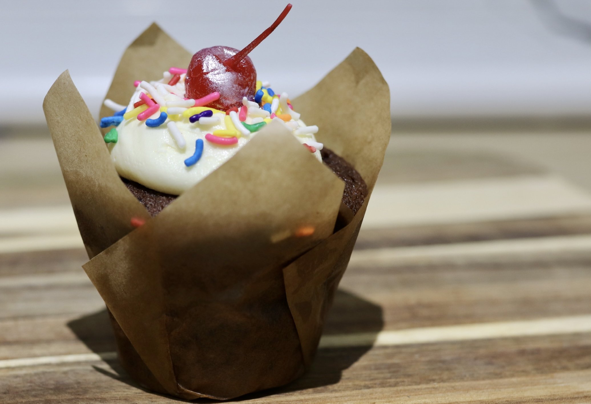 Chocolate cupcake with frosting, sprinkles, and a cherry on top in a paper liner on a wooden surface.