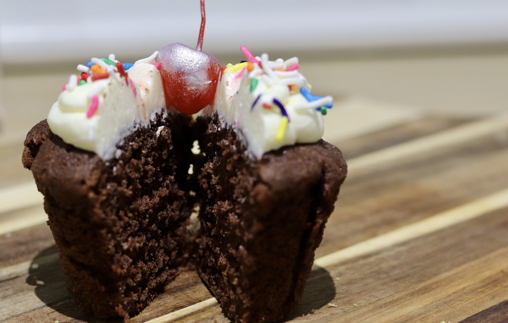 Chocolate cupcake cut in half, topped with white frosting, colored sprinkles, and a cherry, on a wooden surface.