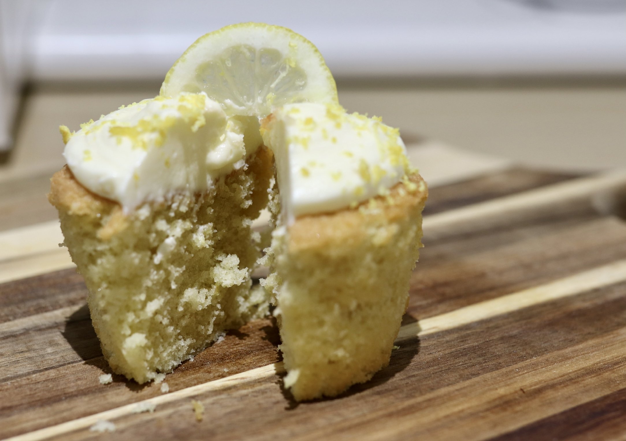 Lemon cupcake with frosting and lemon zest, topped with a lemon slice, cut in half on a wooden board.