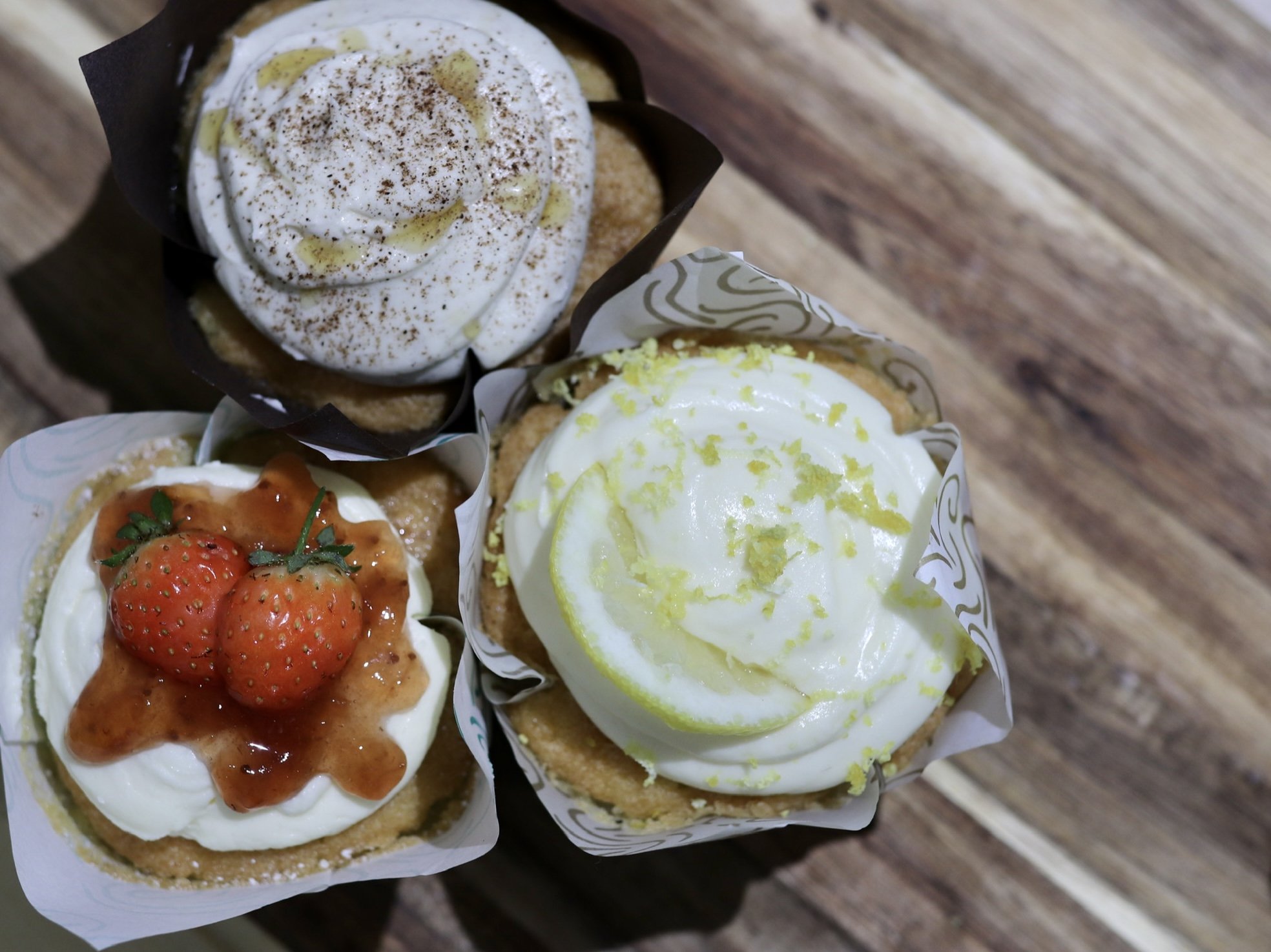Three cupcakes with different toppings: one with whipped cream and cinnamon, one with strawberries and syrup, and one with lemon zest on icing, on a wooden surface.