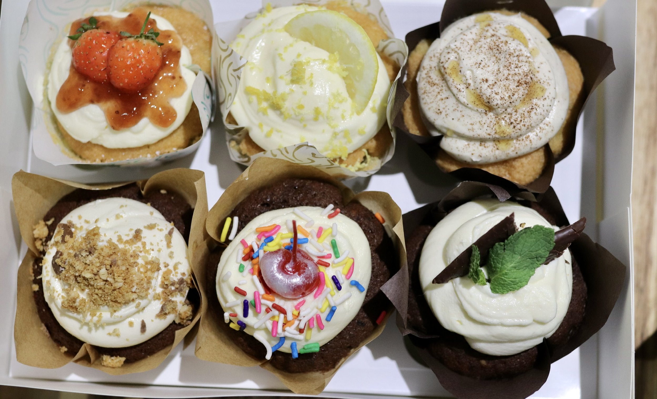 Assorted decorated cupcakes with various toppings such as strawberries, lemon slice, cinnamon, crumbled nuts, sprinkles, cherry, chocolate piece, and mint leaf in a box.