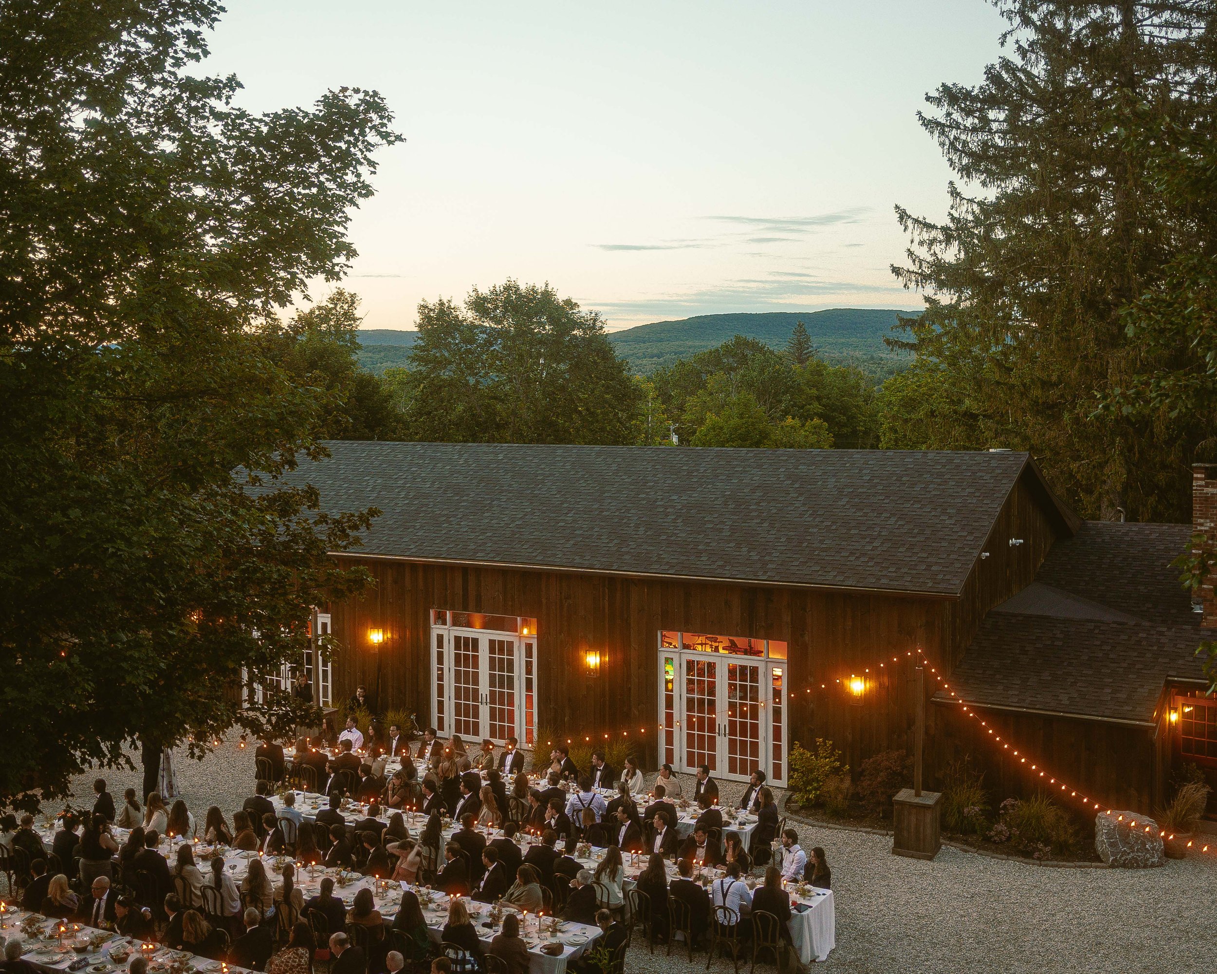 An outdoor evening wedding reception with guests seated at long tables with white tablecloths, string lights, and warm lighting, surrounded by trees and a rustic building, with a scenic mountain view in the background.