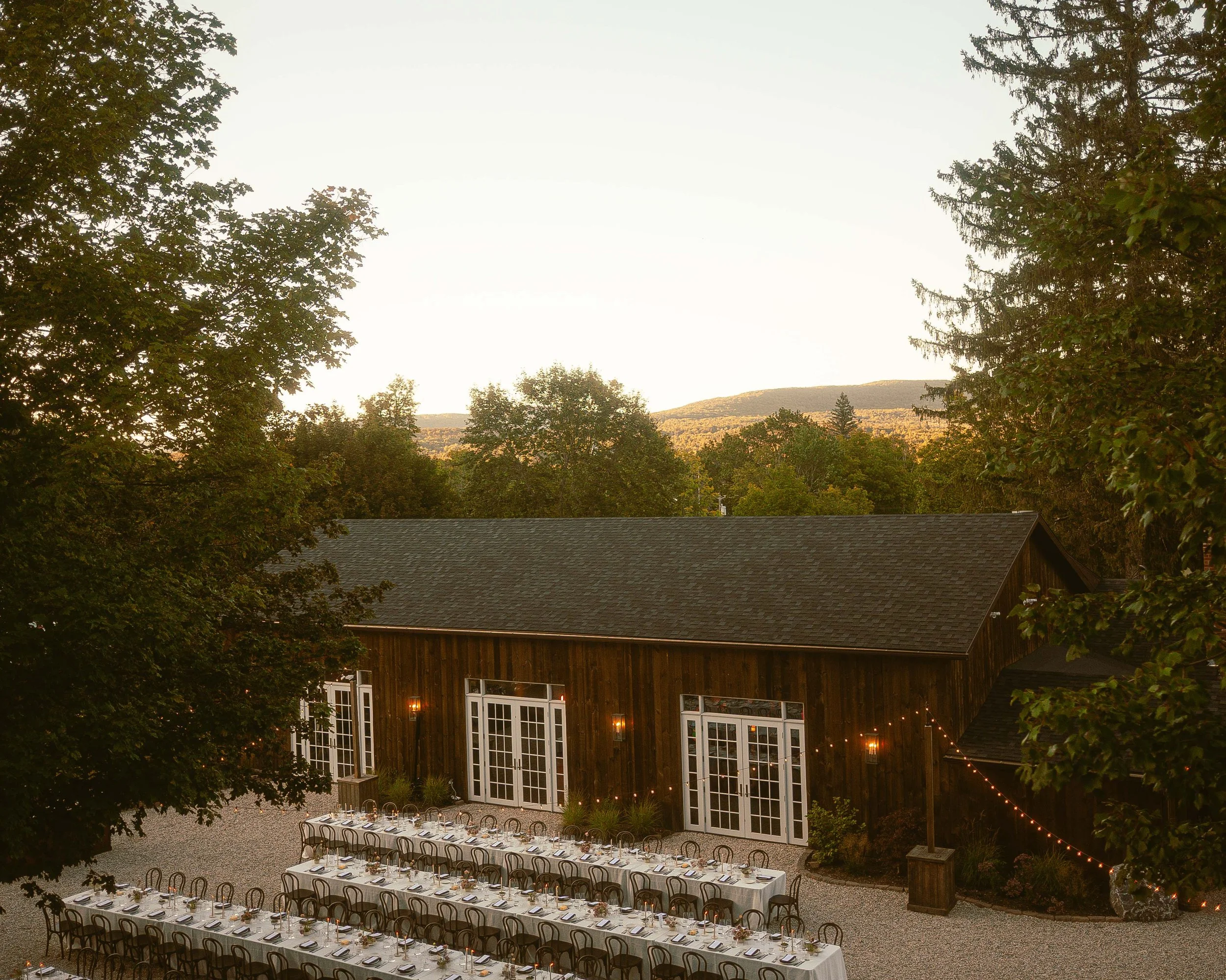 An outdoor wedding reception setup at sunset with a long banquet table, chairs, and a rustic barn, surrounded by trees and mountain views.