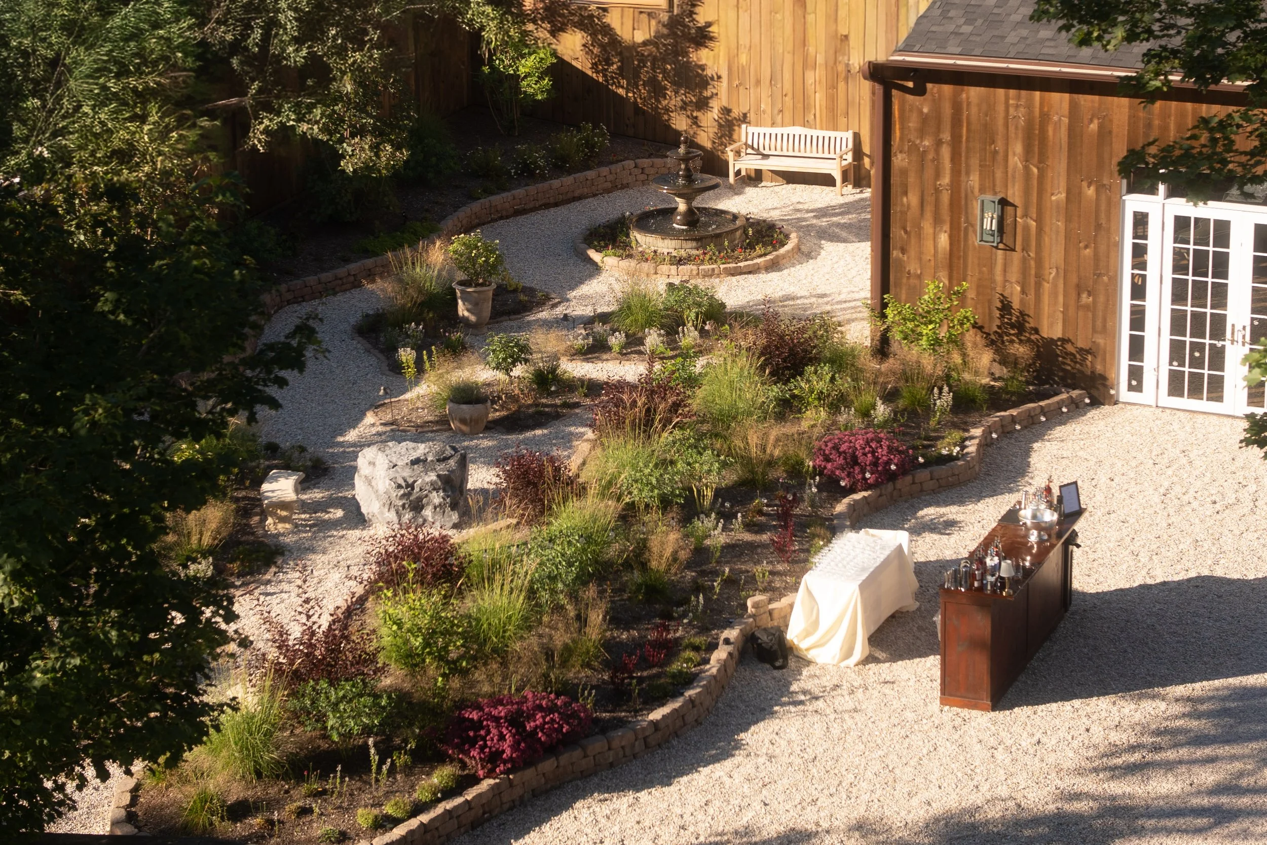 A backyard garden with a gravel pathway, a decorative fountain in the center, and various plants and flowers. There is a wooden fence, a pink bench, and a house with white French doors and outdoor lighting.