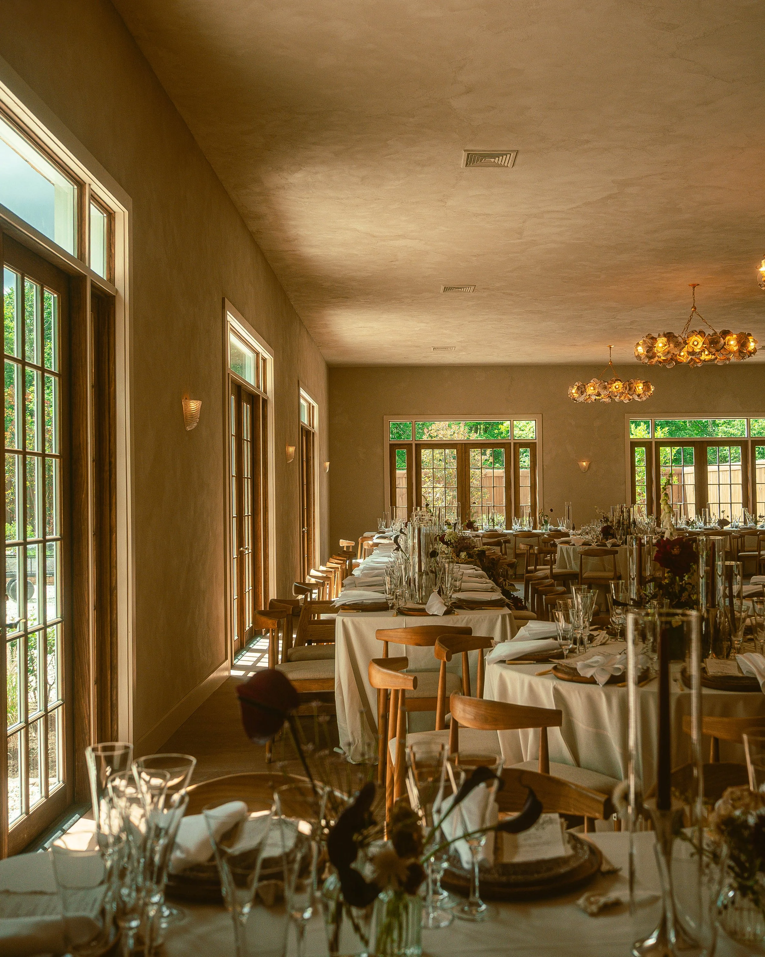 Elegant indoor dining area with large windows, round chandeliers, and a long set table with white tablecloths, glassware, and floral centerpieces.