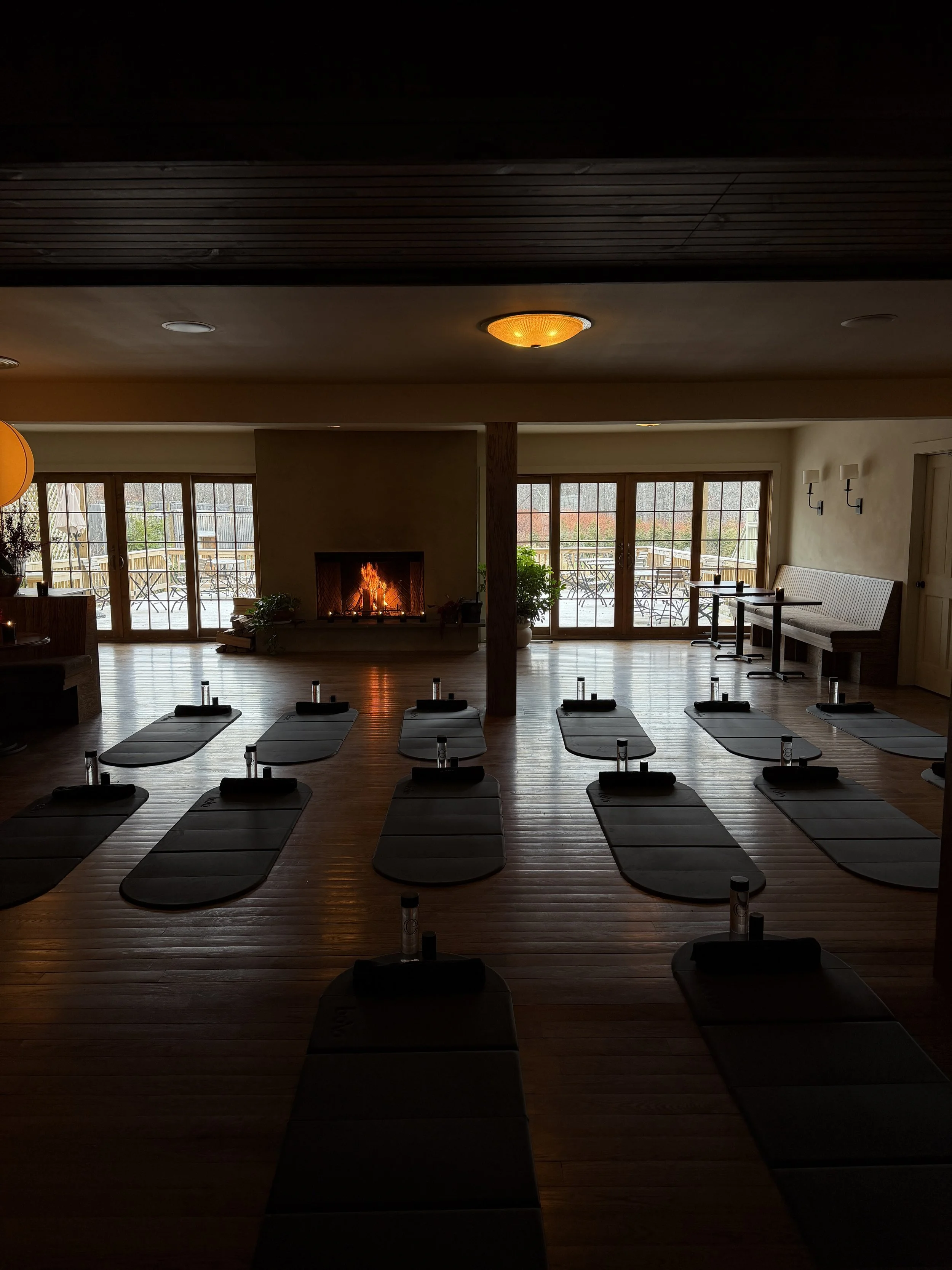 Yoga studio with black mats arranged on wooden floor, fireplace, and large windows with outdoor seating.
