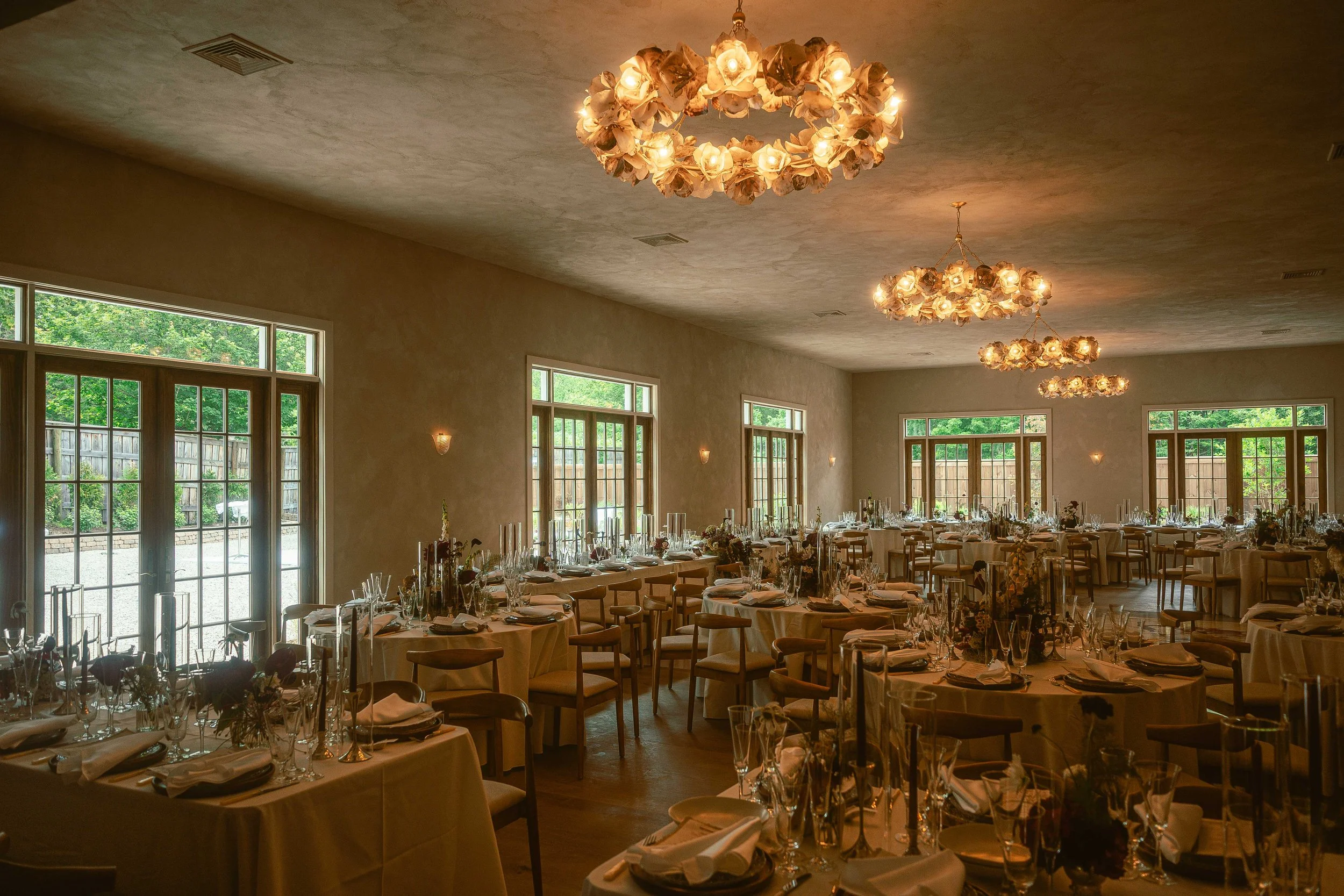 Elegant banquet hall with round tables set with white tablecloths, glassware, and floral centerpieces, large windows with greenery outside, and chandeliers hanging from the ceiling.