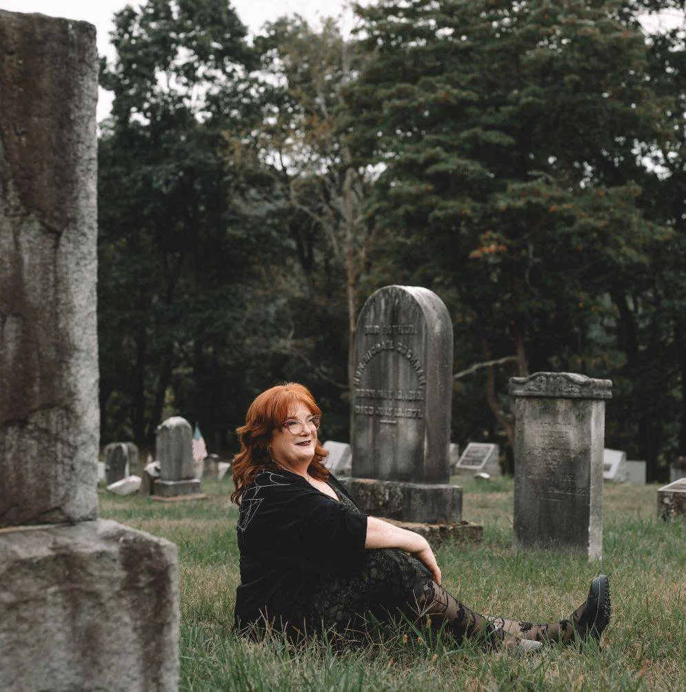 A woman with red hair and glasses sitting on the grass in a cemetery, surrounded by gravestones and trees.