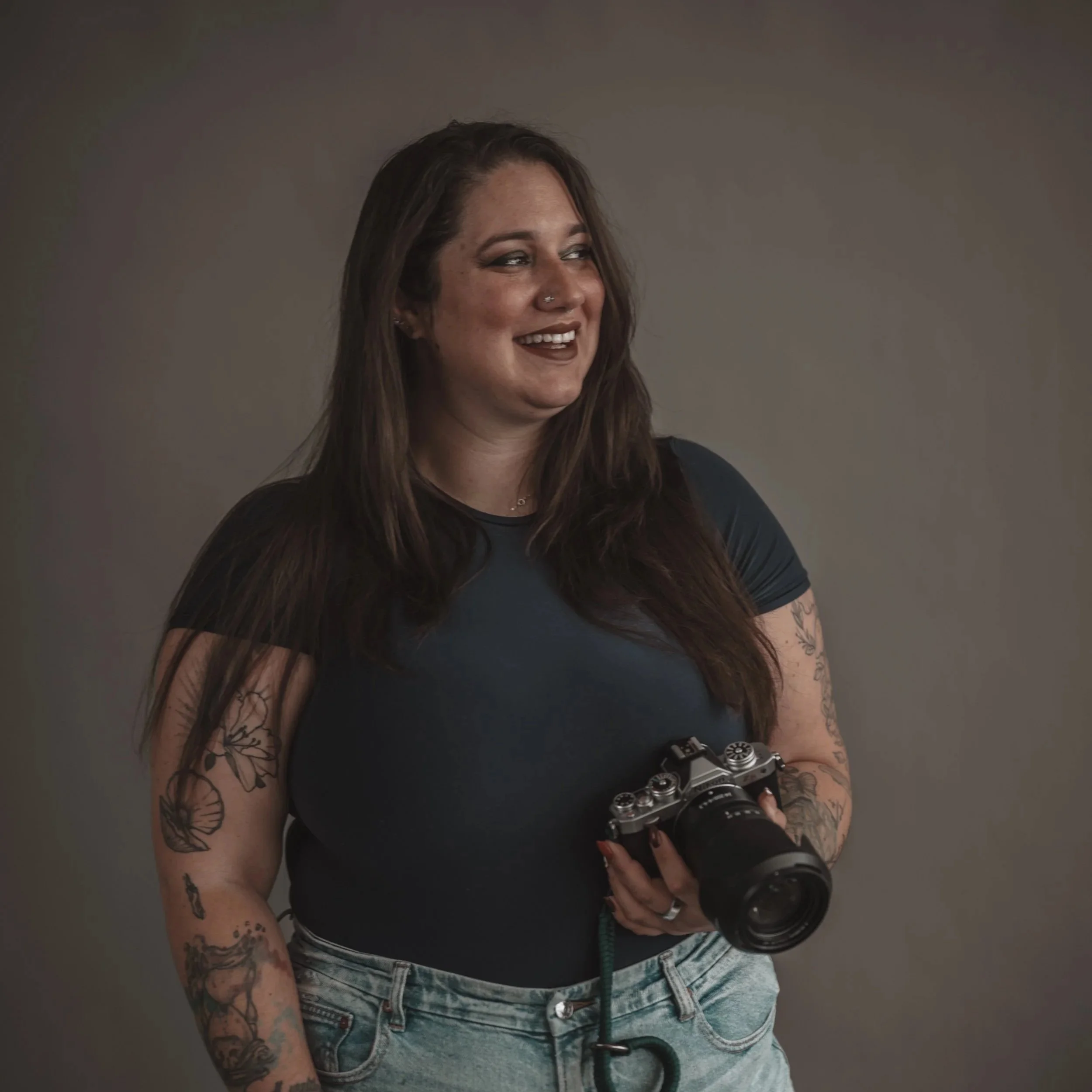 A woman with shoulder-length brown hair and tattoos on both arms is smiling and holding a camera in her right hand. She is wearing a black shirt and light-wash jeans, standing against a plain gray background.