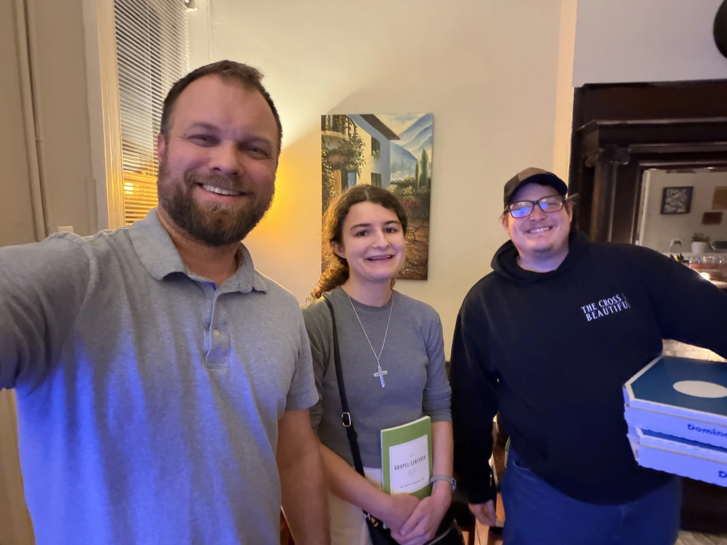 Three smiling people, two men and one woman, standing indoors during a gathering or event. One man has a beard and is wearing a light gray polo shirt, the woman has curly hair and is wearing a gray shirt with a cross necklace, and the other man is we