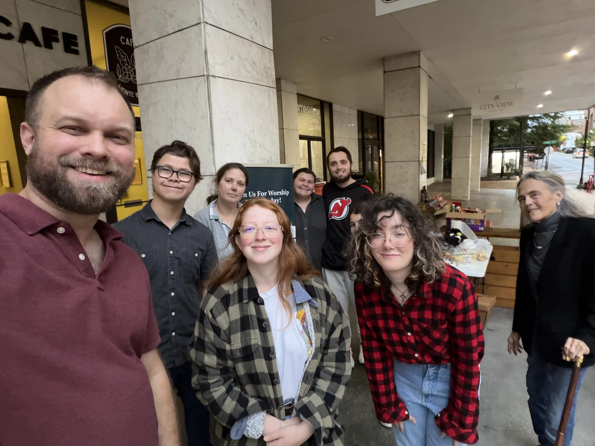 A group of people standing outside a building taking a selfie, with some people smiling and others neutral, with tables of food and a sign in the background.