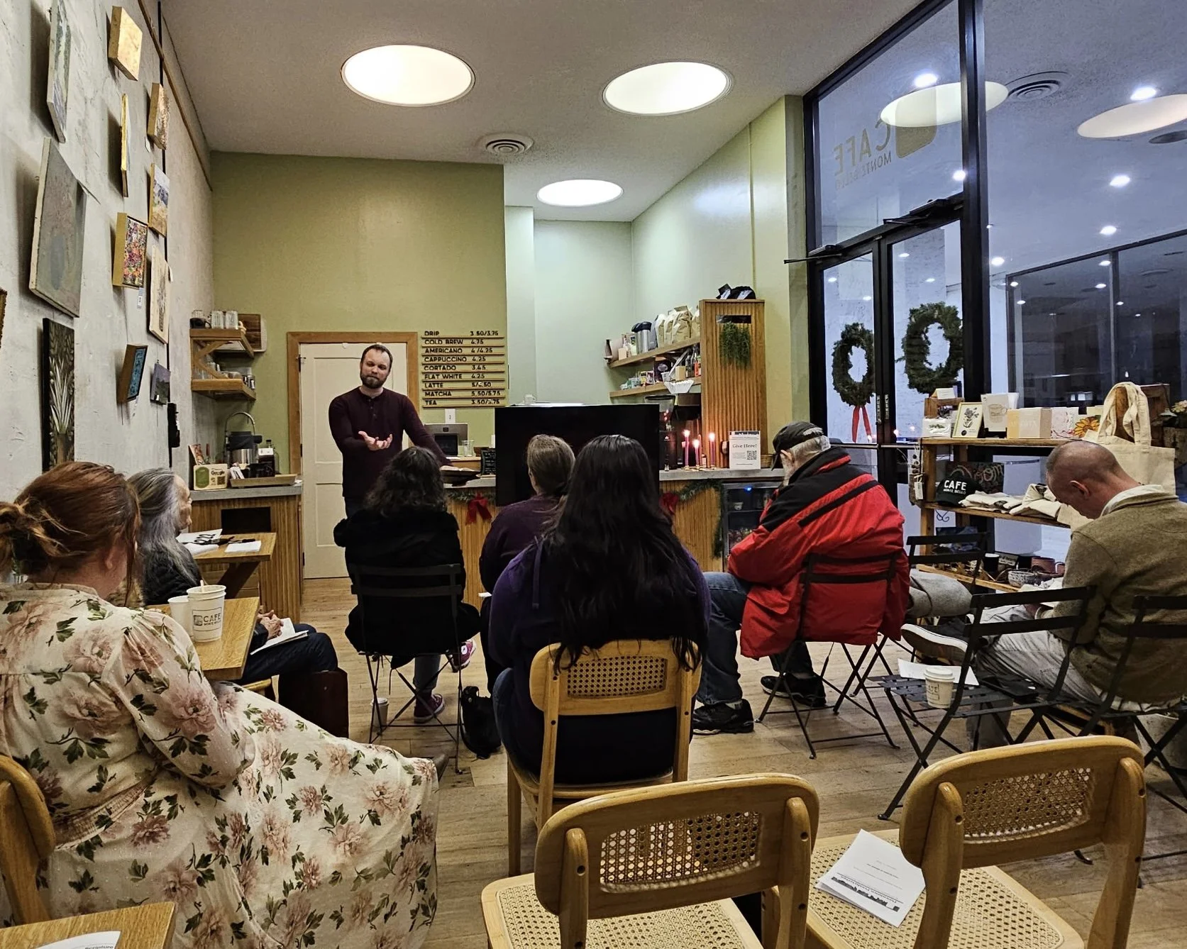 A group of people attending a presentation or meeting in a cozy coffee shop with green walls, wooden furniture, and decorative art. The presenter stands near a counter, speaking to the audience seated on chairs and benches around. Giant windows show 