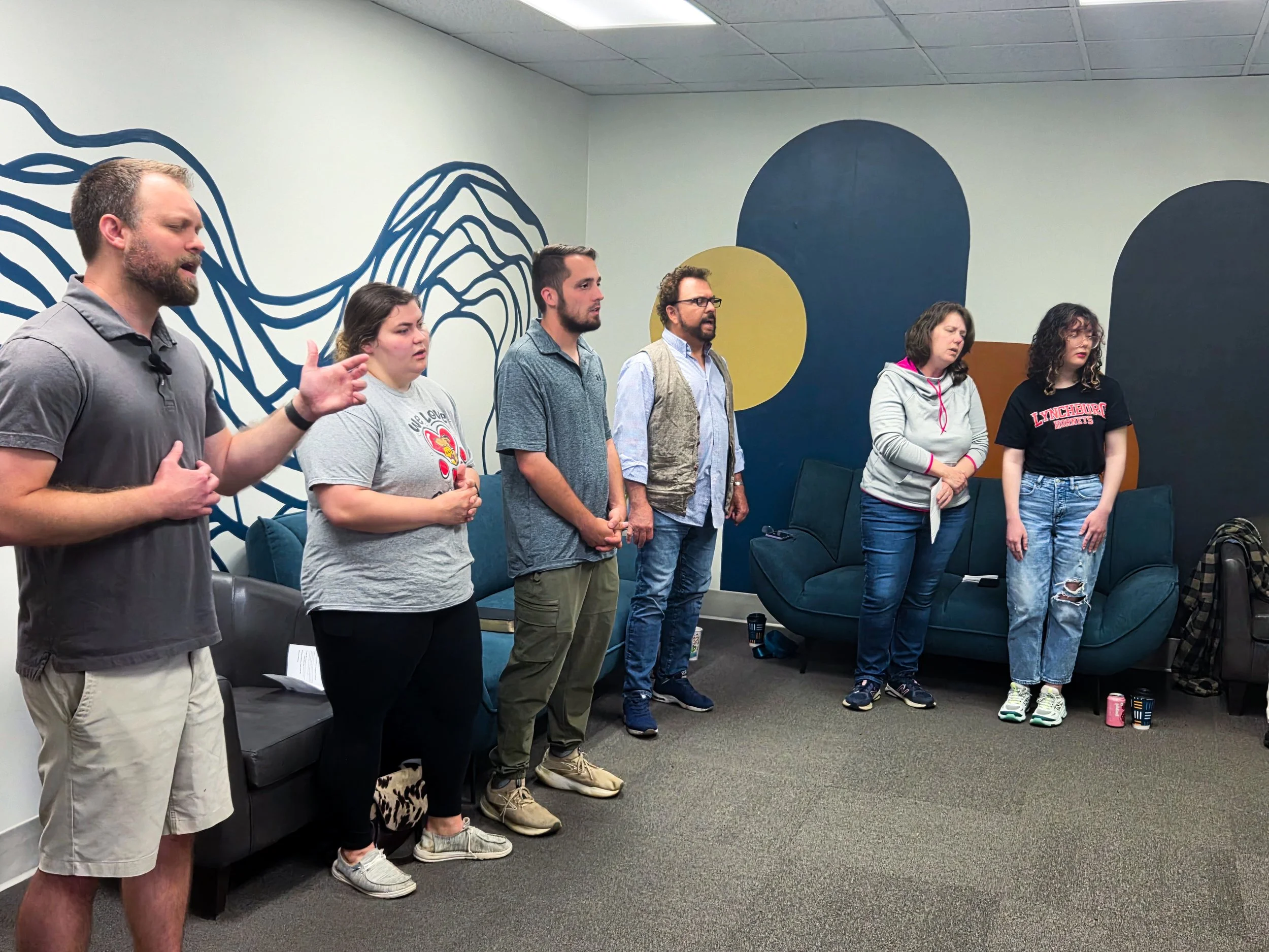 A group of six people standing side by side in a room with a mural on the wall, appearing engaged in a discussion or listening intently.