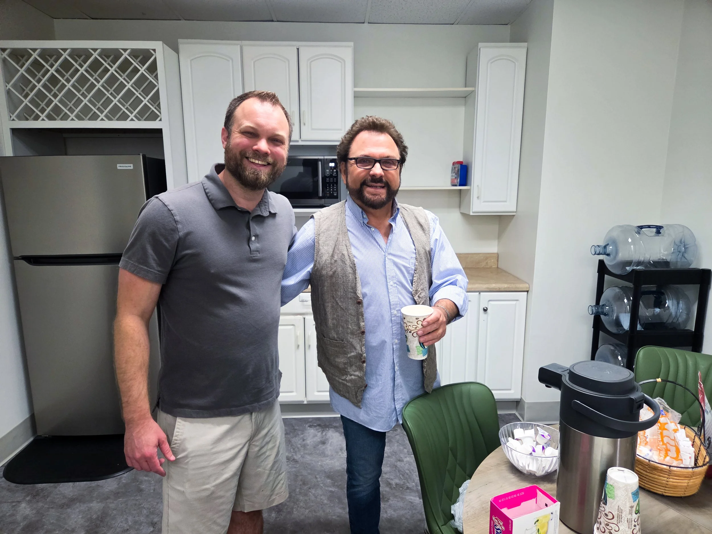 Two men standing in a kitchen, smiling at the camera. One is wearing a gray polo shirt, and the other is wearing glasses, a blue shirt, and a vest. They are holding cups. The kitchen has white cabinets, a microwave, and a refrigerator. There are chai
