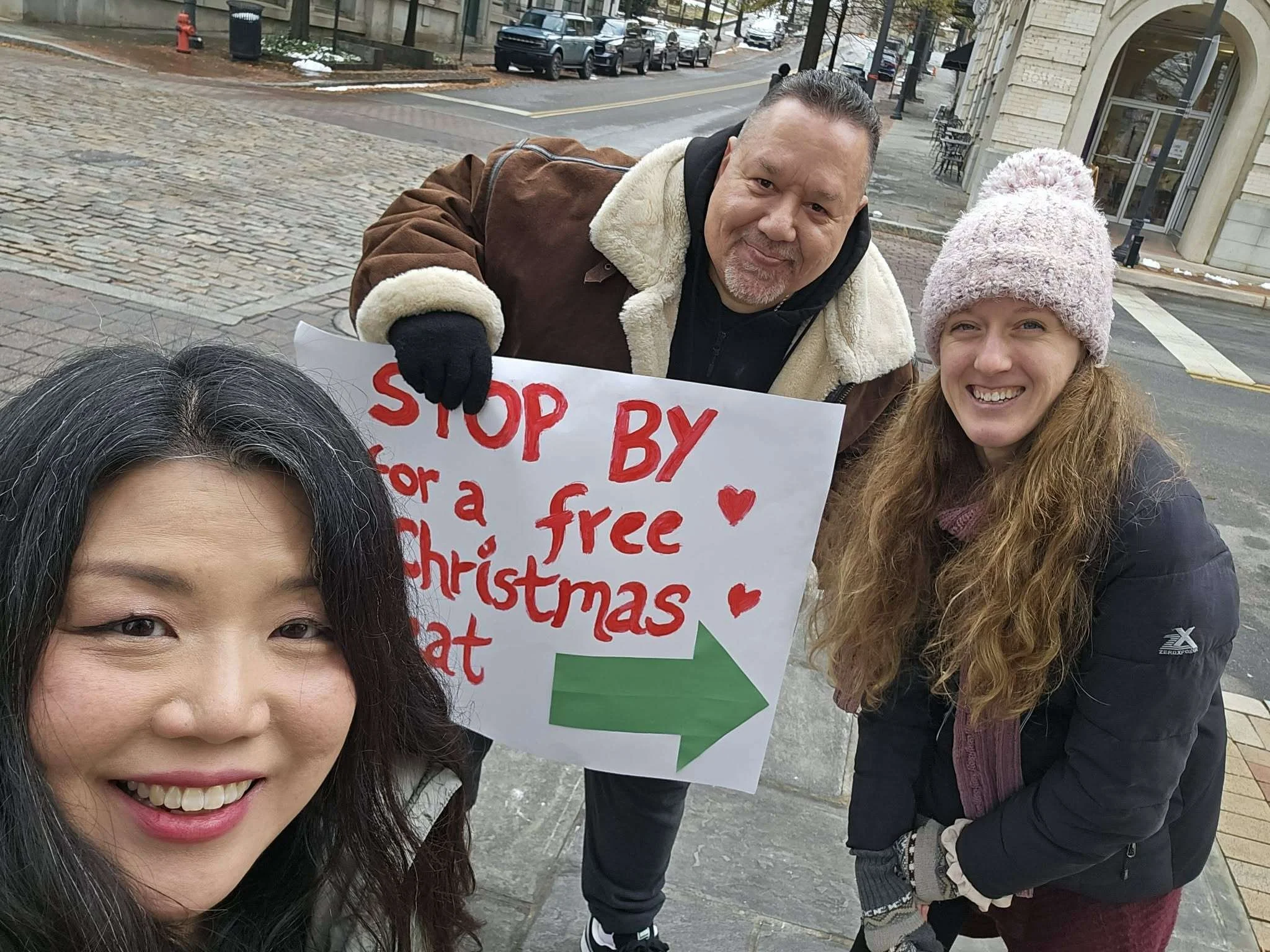 Group of people standing on a city sidewalk, holding a sign that says 'Stop by for a free Christmas' with a green arrow pointing to the right. They are smiling and wearing winter clothing, including a pink knit hat, a black jacket, and a brown jacket