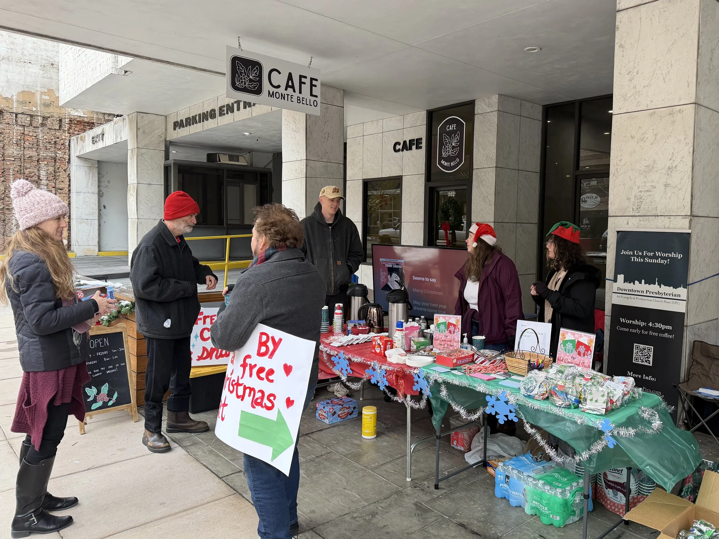 People shopping at a Christmas charity event outside a cafe, with a table of holiday items, signs, and festive decorations.