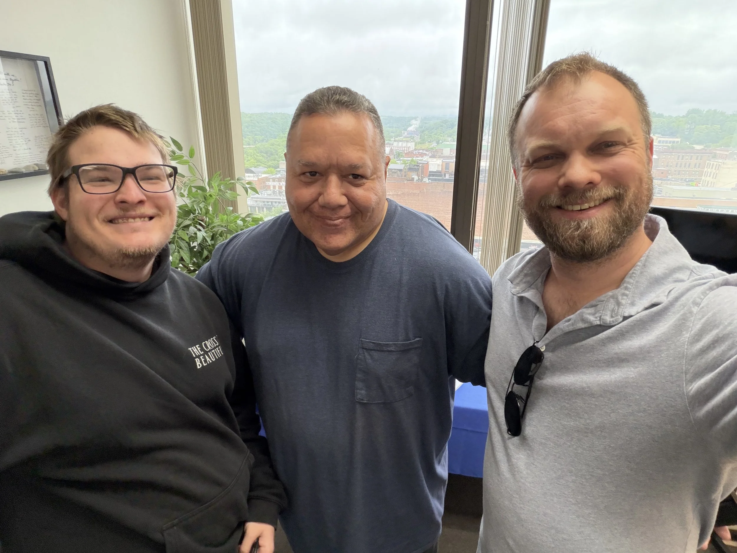 Three men smiling for a photo indoors, with large windows showing a cityscape and green hills in the background.