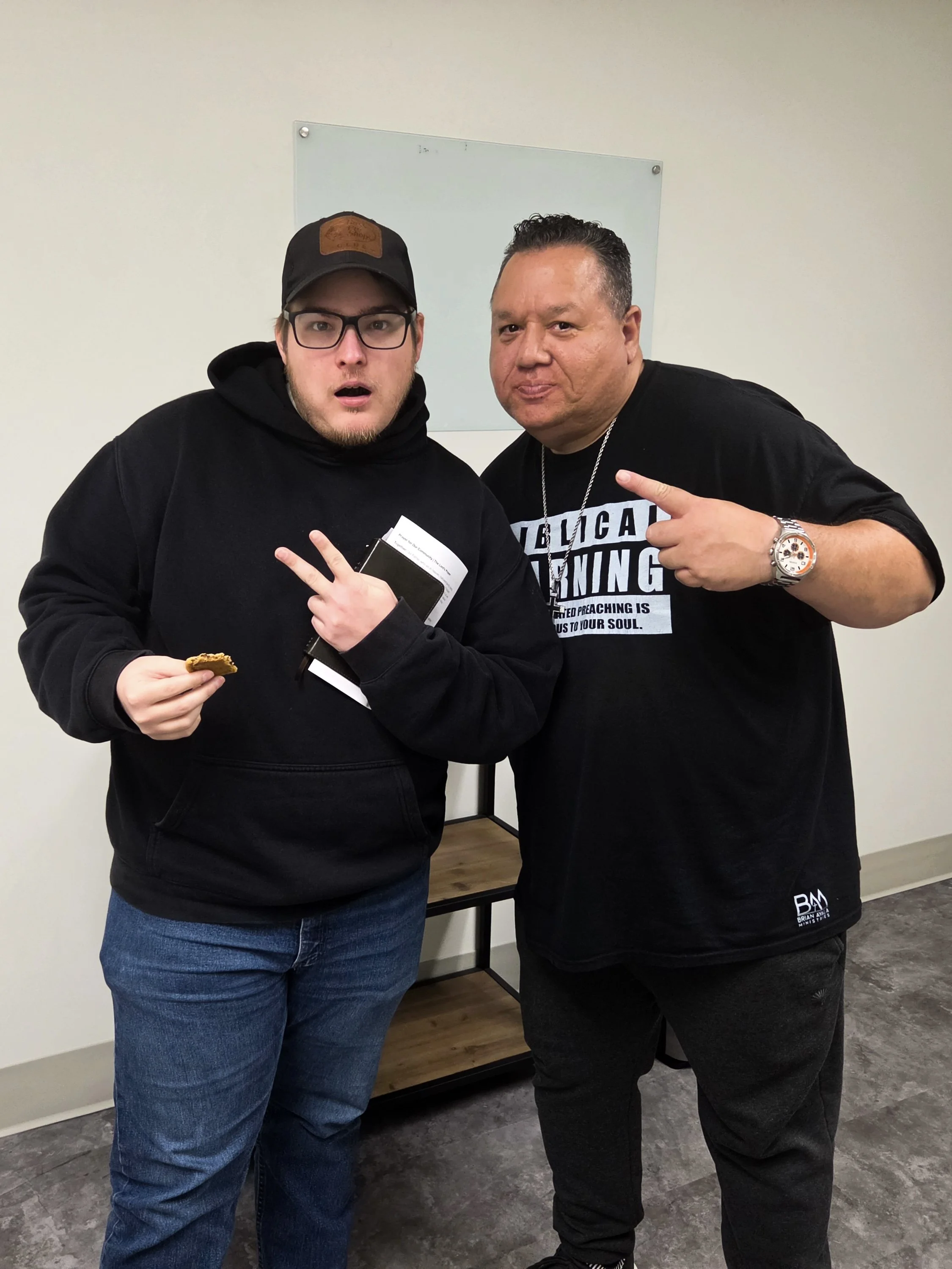 Two men standing close together indoors against a plain wall, both making peace signs with their hands and looking at the camera. The man on the left is wearing glasses, a black hoodie, and a black cap, holding a snack and some papers. The man on the