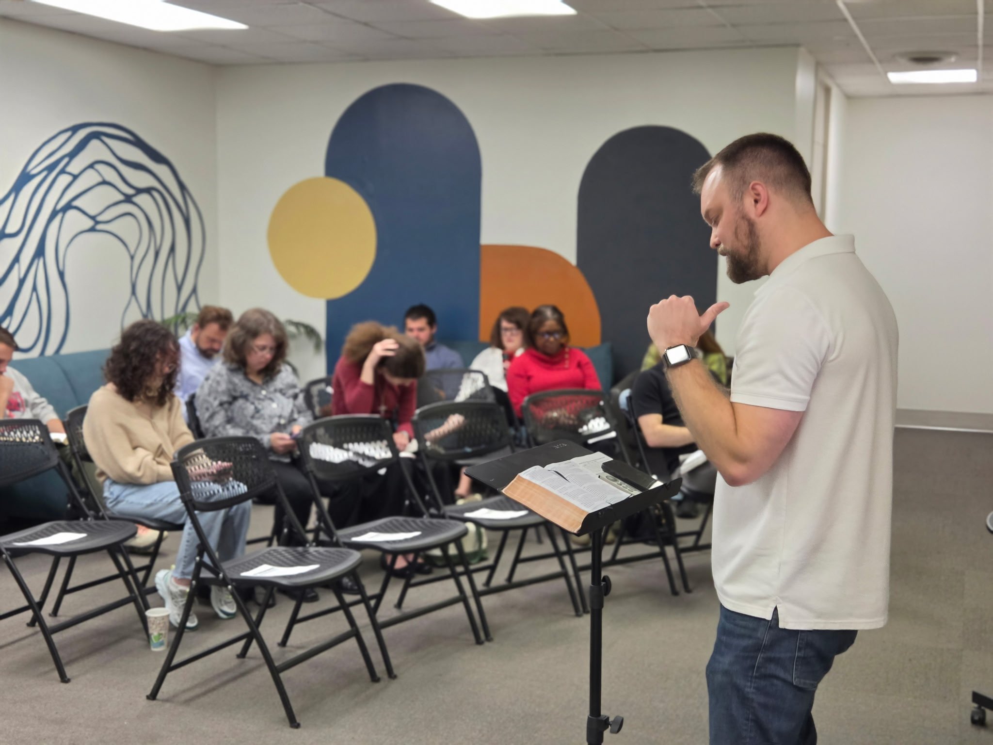 A man with a beard wearing a white shirt and smartwatch is standing in front of a group of people seated in black chairs. The group is in a room with modern decor, including a wall with abstract mural art and ceiling lights.