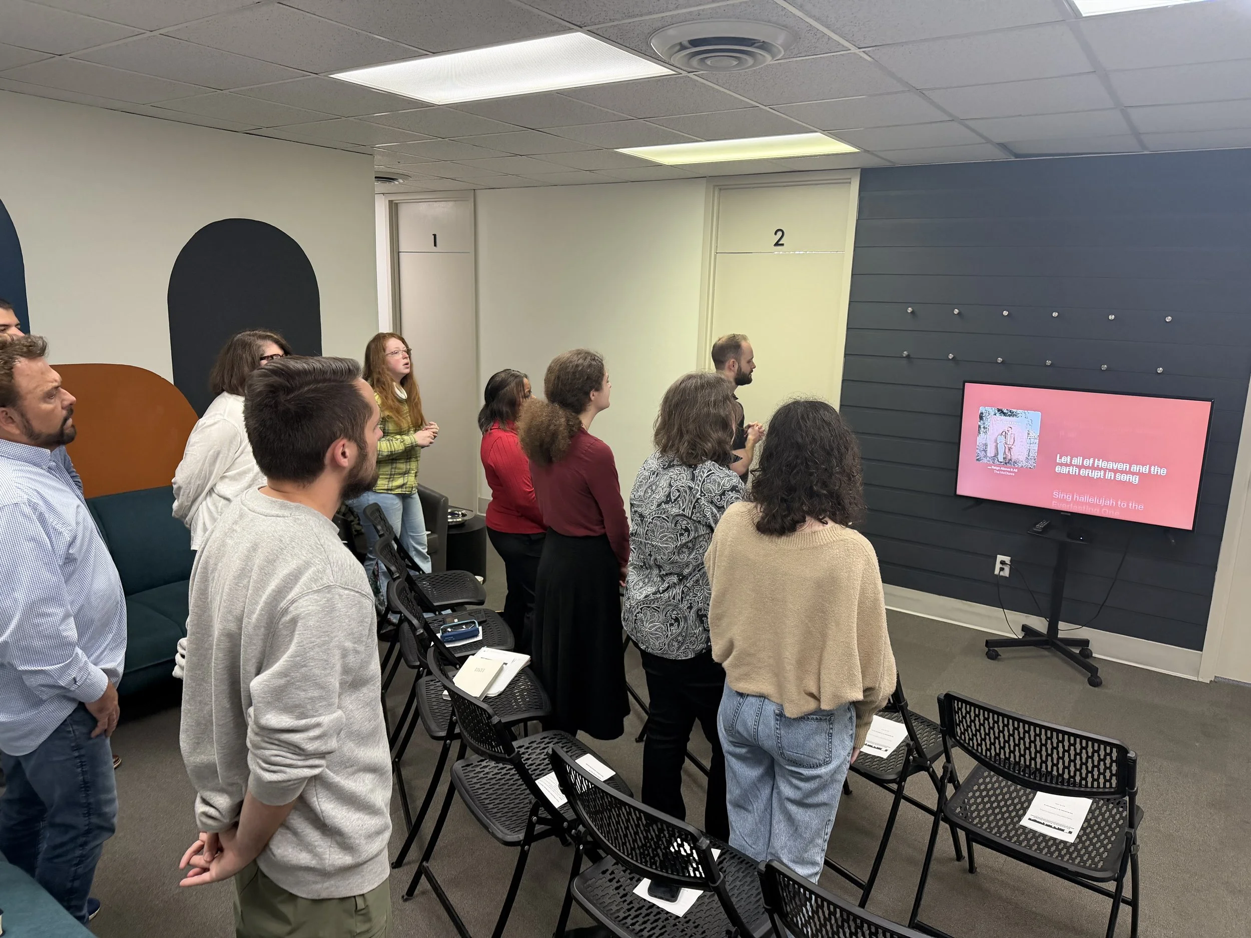 A group of people standing in a room, facing a large screen displaying song lyrics, while some sit on chairs. The room has a white ceiling with fluorescent lights and a dark blue accent wall with hooks. The audience appears engaged, possibly singing 