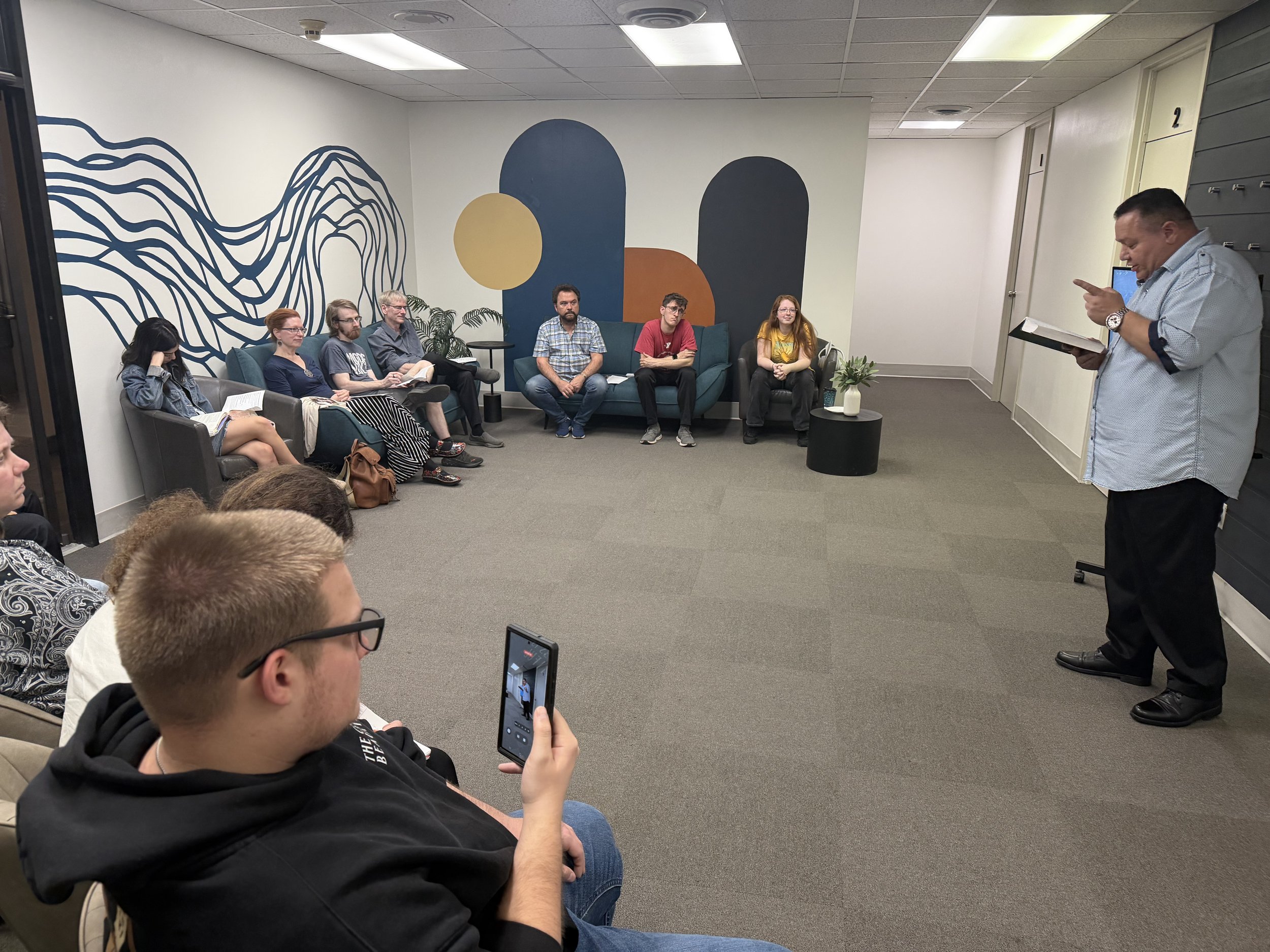 A group of people seated on couches and chairs in a room with colorful wall art, listening to a man standing and reading from a book. A person in the foreground is taking a photo or recording with a tablet.