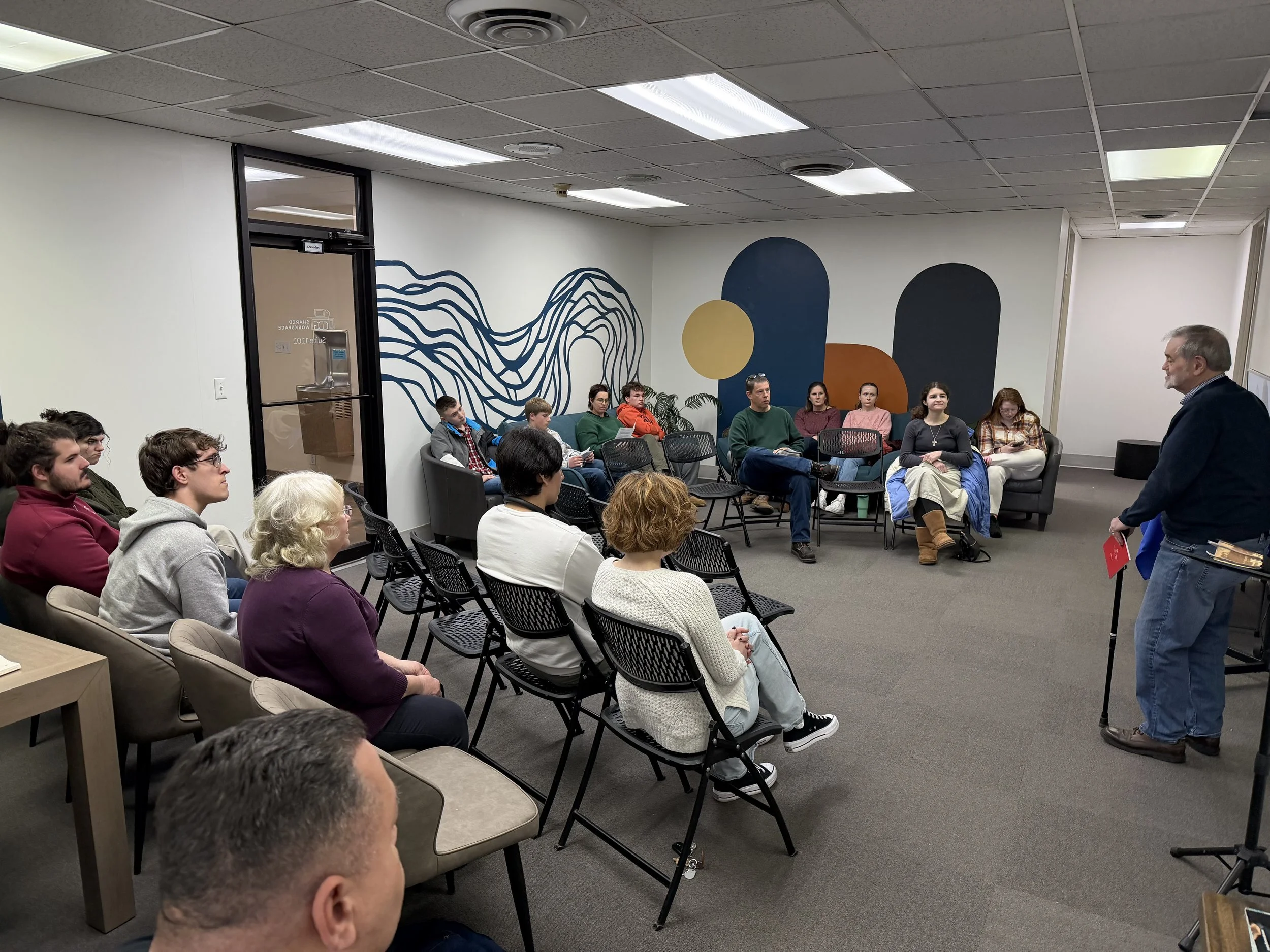 A man is standing and speaking to a group of seated people in a conference room with abstract wall art.