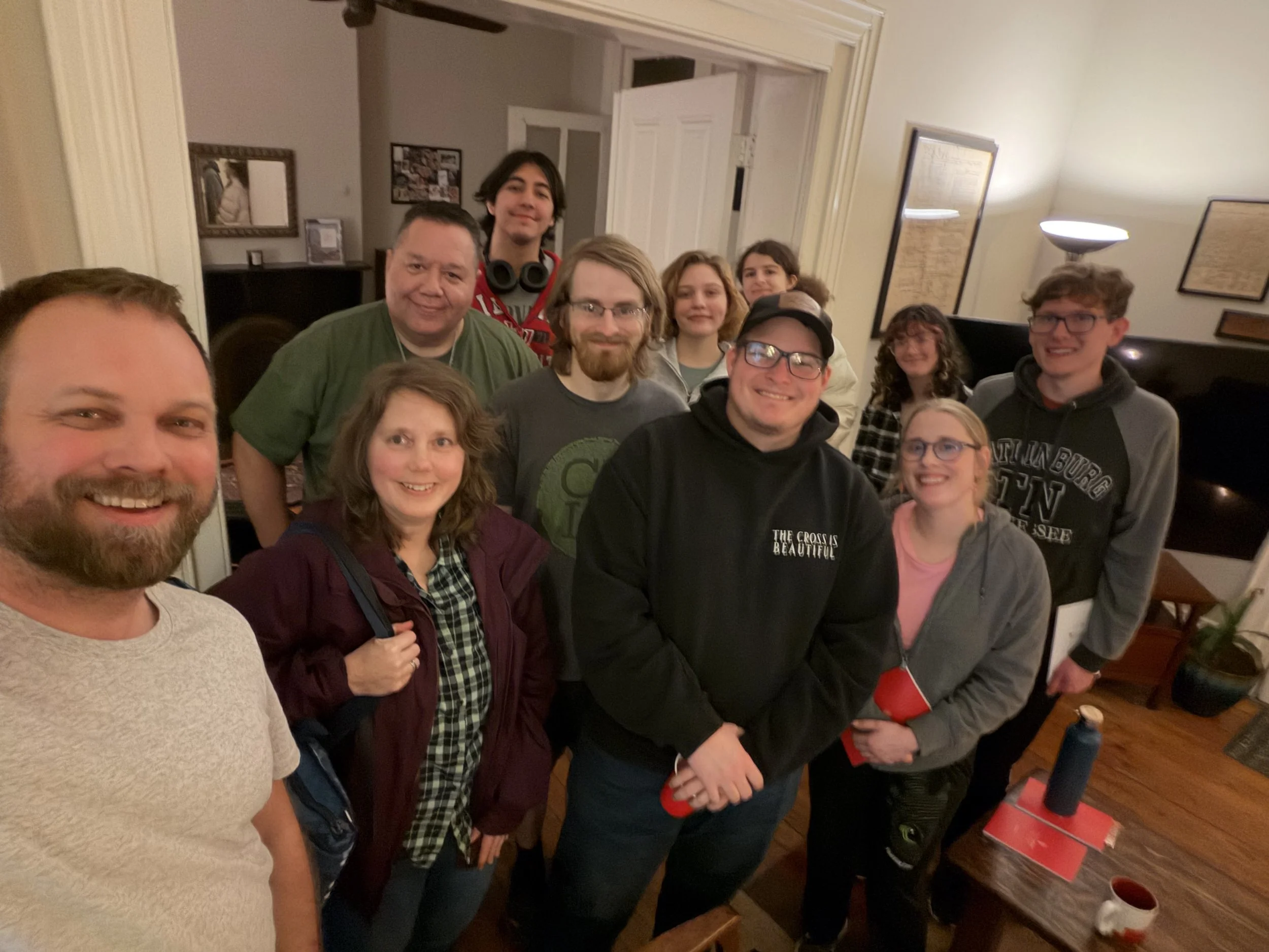 A group of people gathered indoors, posing for a photo; some are smiling and looking at the camera, while others are looking elsewhere. The room has framed pictures and a lamp on the wall, with a table containing a red box and other items.