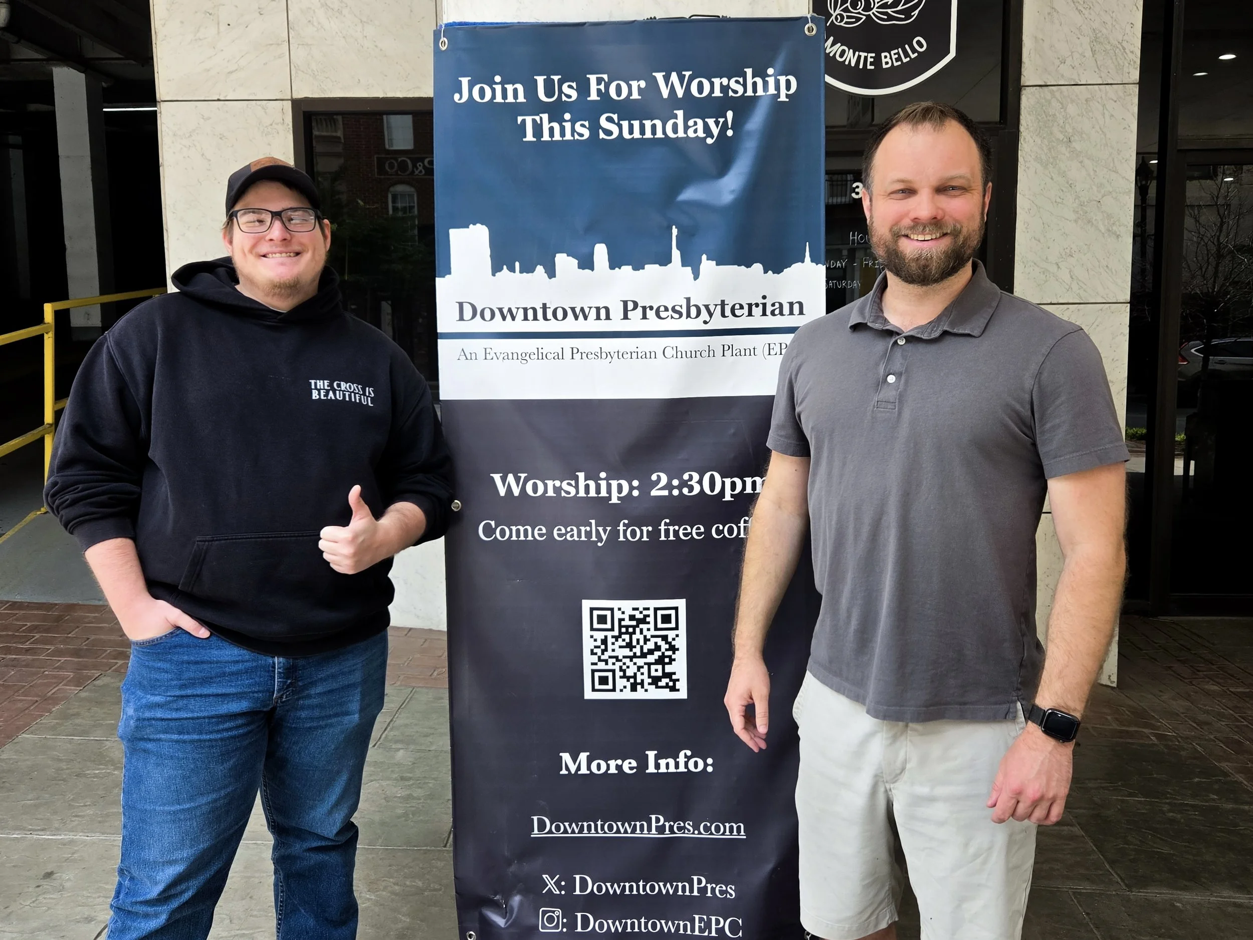 Two men smiling and standing outside next to a blue and black vertical banner for Downtown Presbyterian church, which has a silhouette of a city skyline and QR code. The man on the left is wearing glasses, a black hoodie with white text, and blue jea
