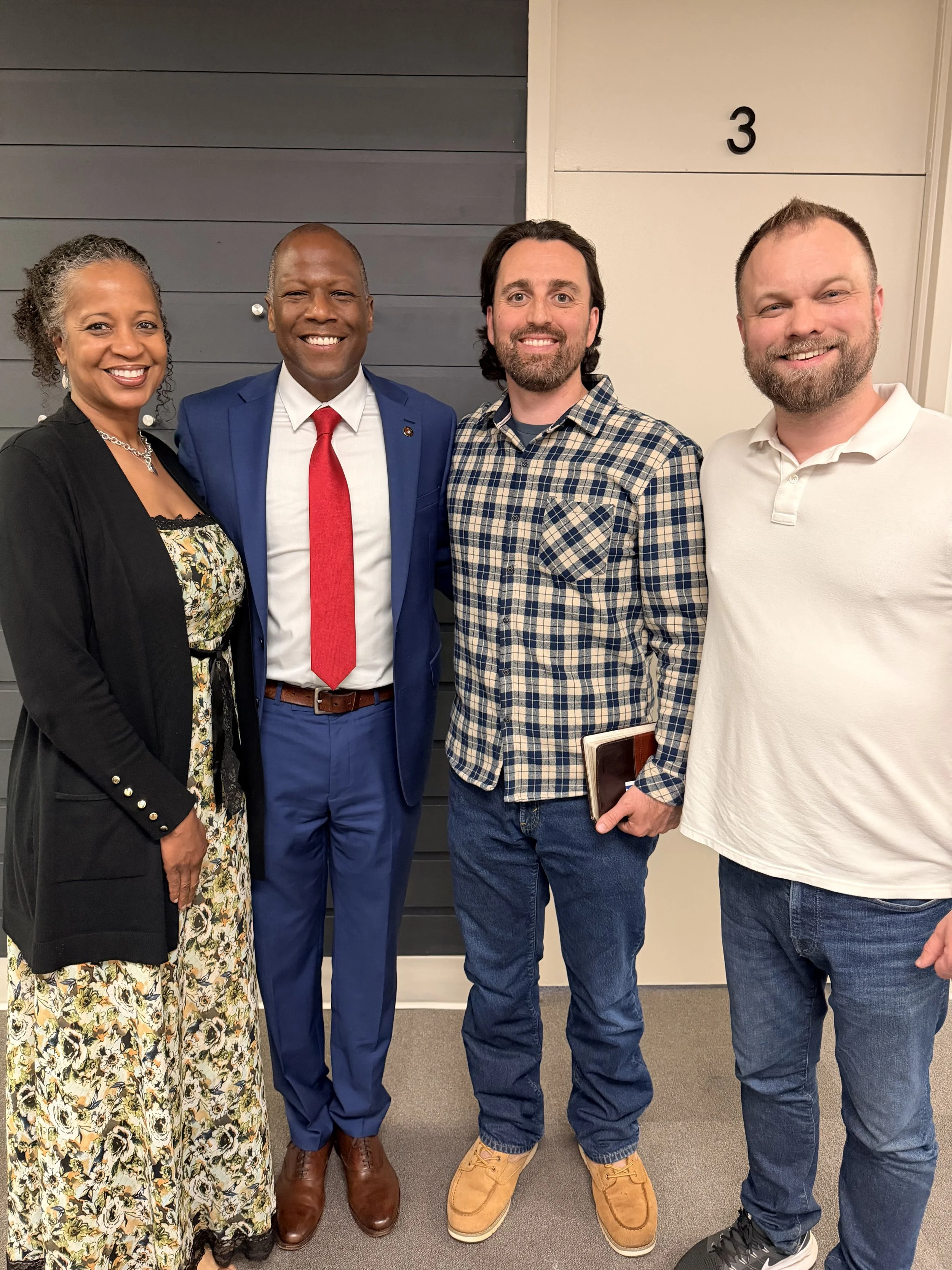 Four diverse adults standing together indoors, smiling at the camera, with a gray and beige wall, and the number 3 visible behind them.