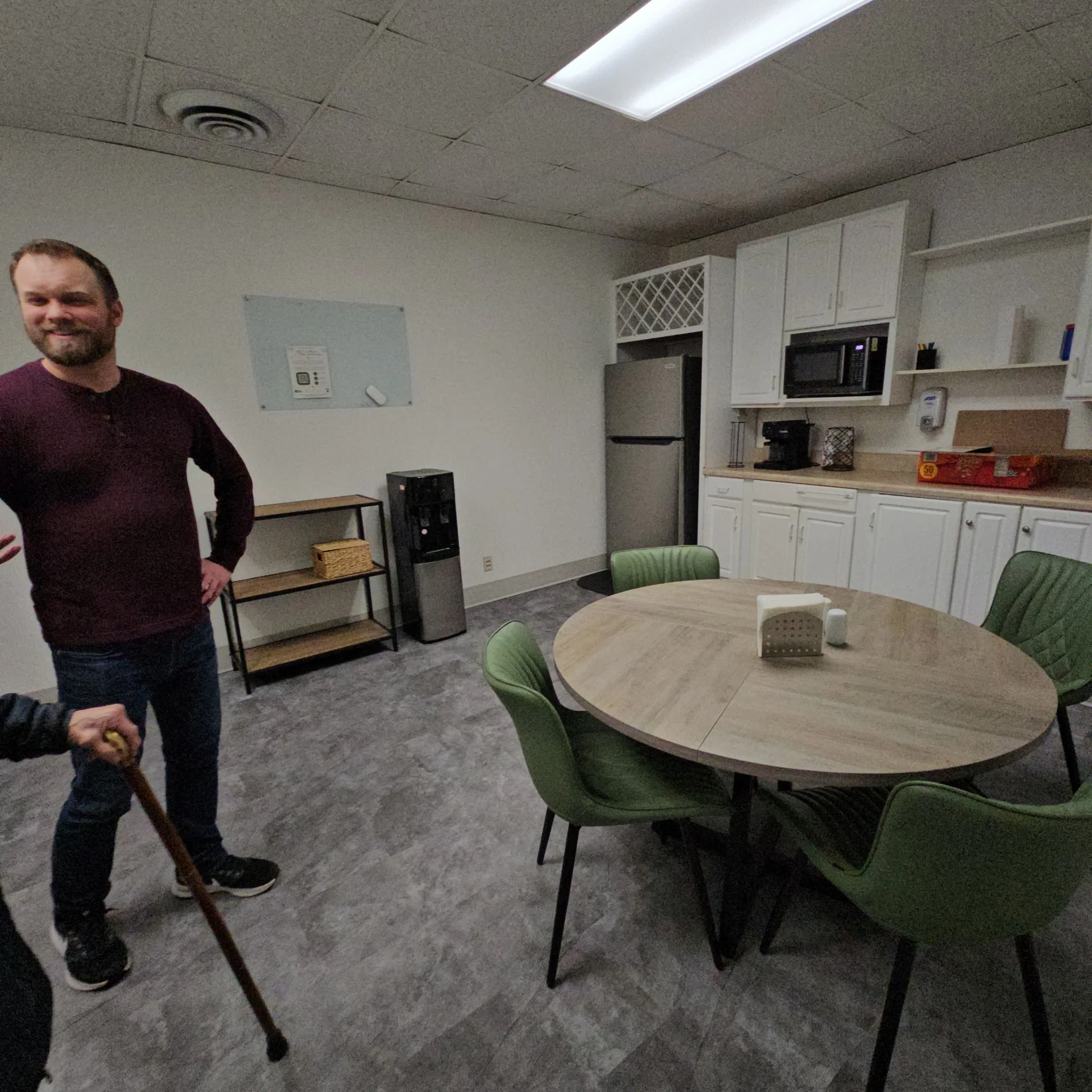 Part of a person standing in a room with a round table and green chairs, kitchen cabinets, microwave, and appliances in the background.