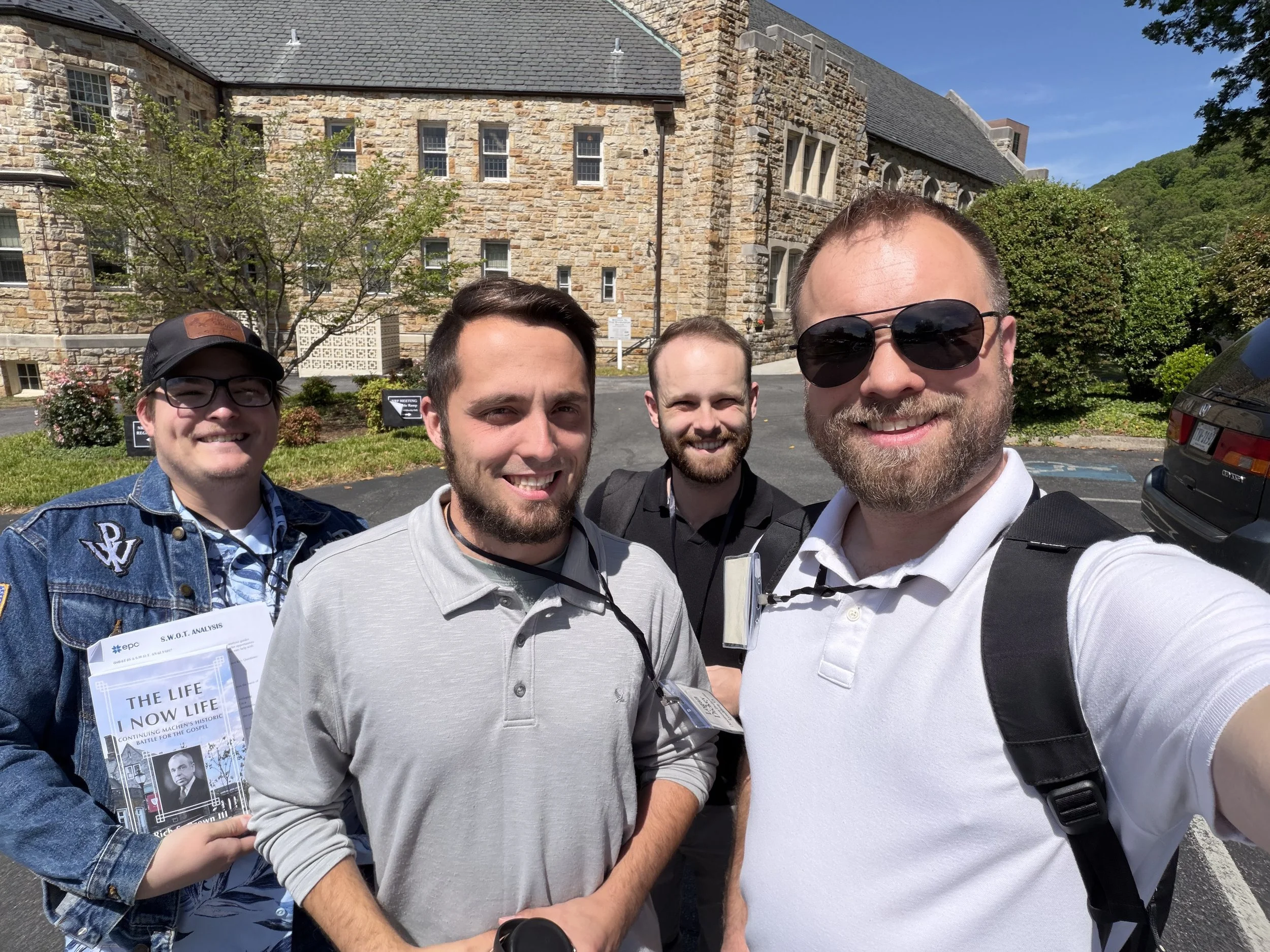 Four men posing for a selfie outside a stone building on a sunny day. One man on the right is wearing sunglasses and a white polo shirt. The others are smiling, with one holding a pamphlet titled 'The Life I Now Life'.
