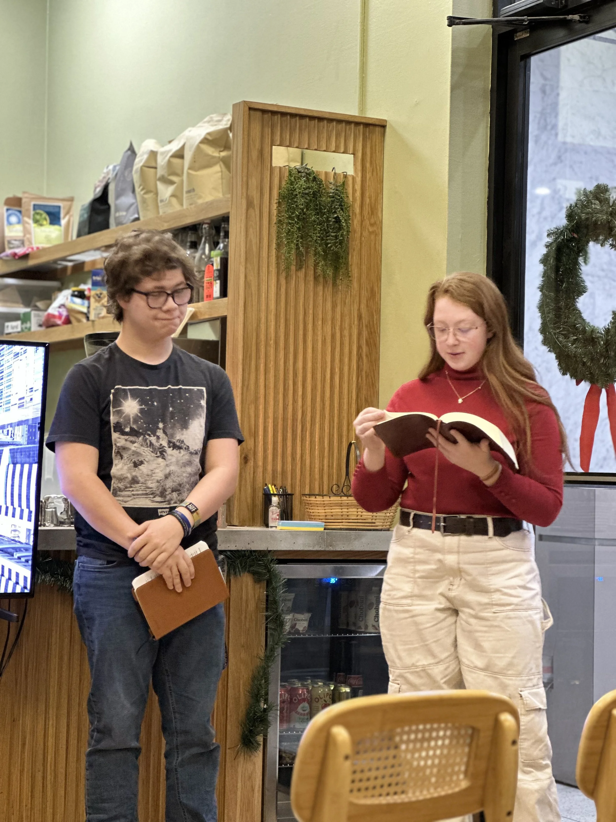 Two young women standing in a cafe, one reading a book and the other holding a notebook, with a wooden and green decor background.