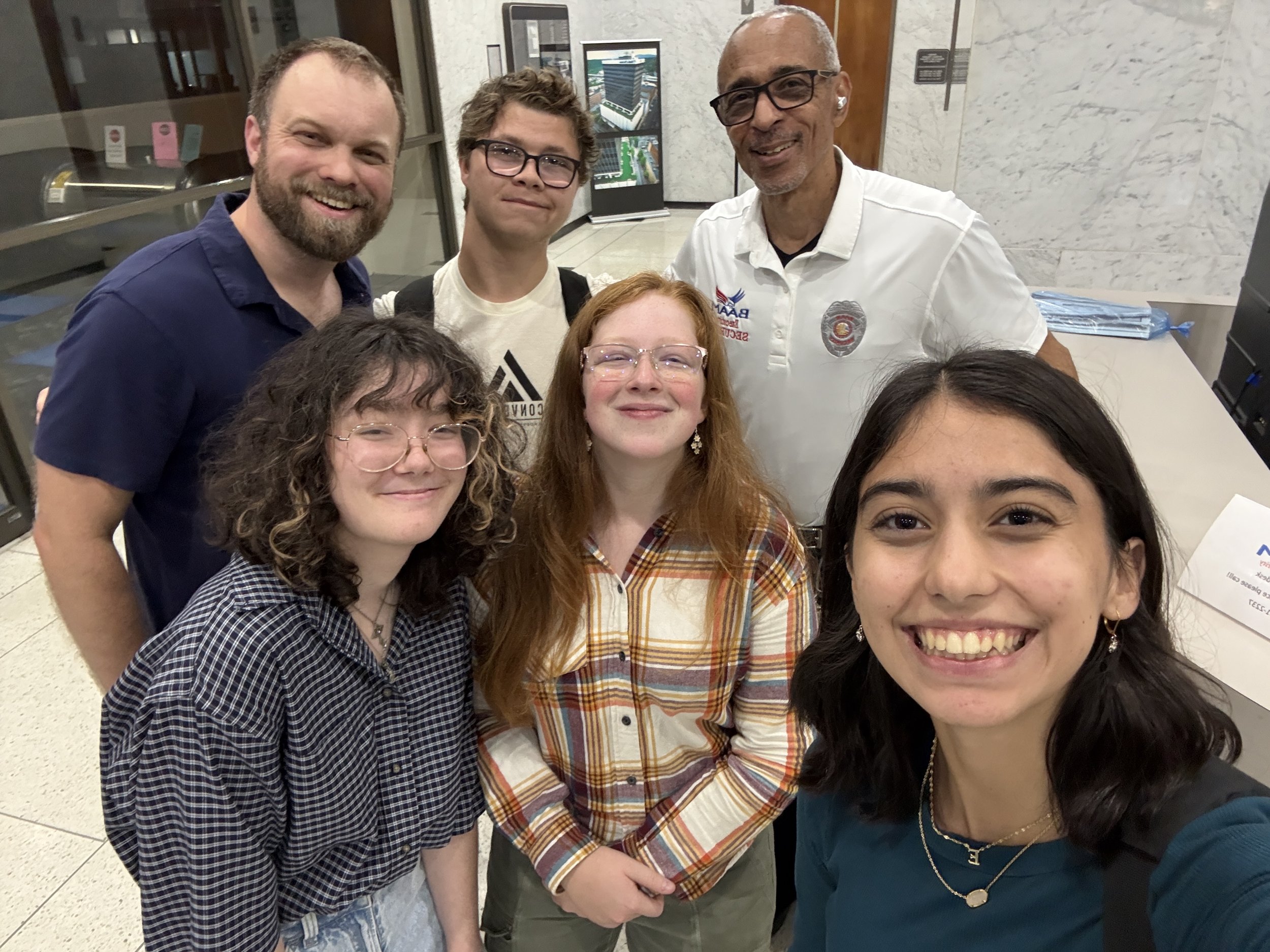 Group of six people taking a selfie indoors, smiling and posing together, with a marble wall and digital displays in the background.