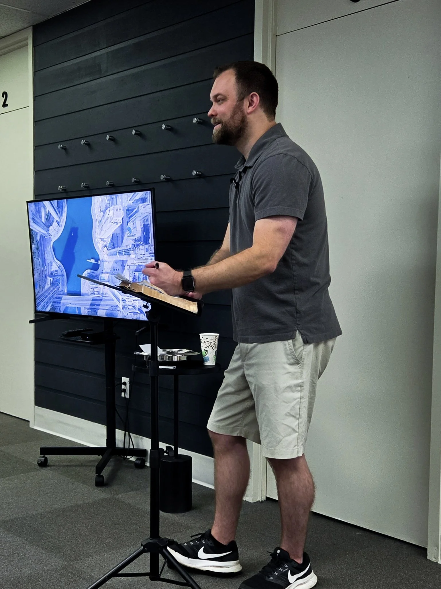 A man with a beard wearing a gray polo shirt, white shorts, and black Nike sneakers standing in front of a display monitor, holding a clipboard and a pen, in an indoor setting with gray carpet and a black wall with hooks.
