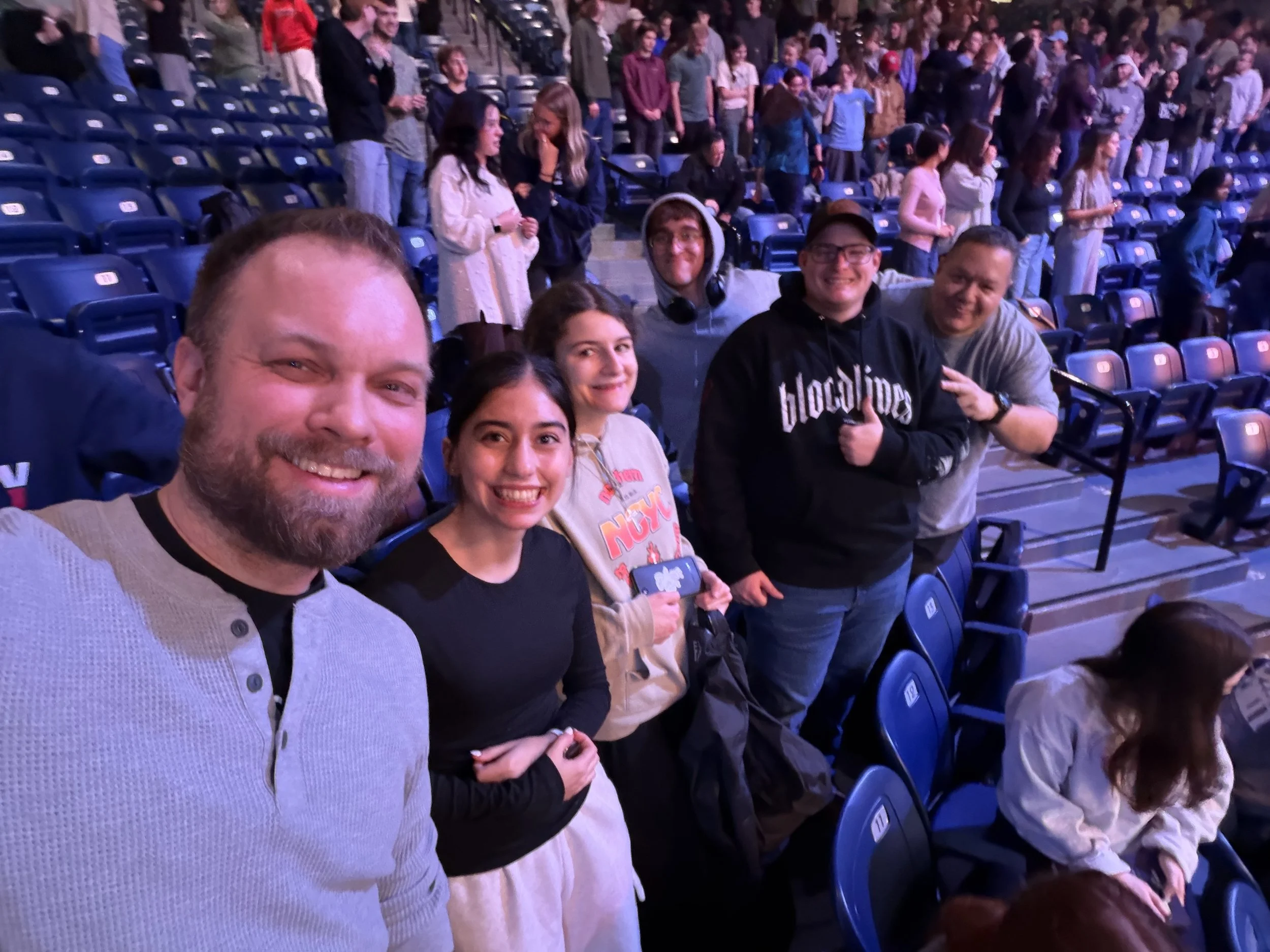 Group of people sitting and standing in an arena, smiling for a selfie, with many empty blue seats in the background.
