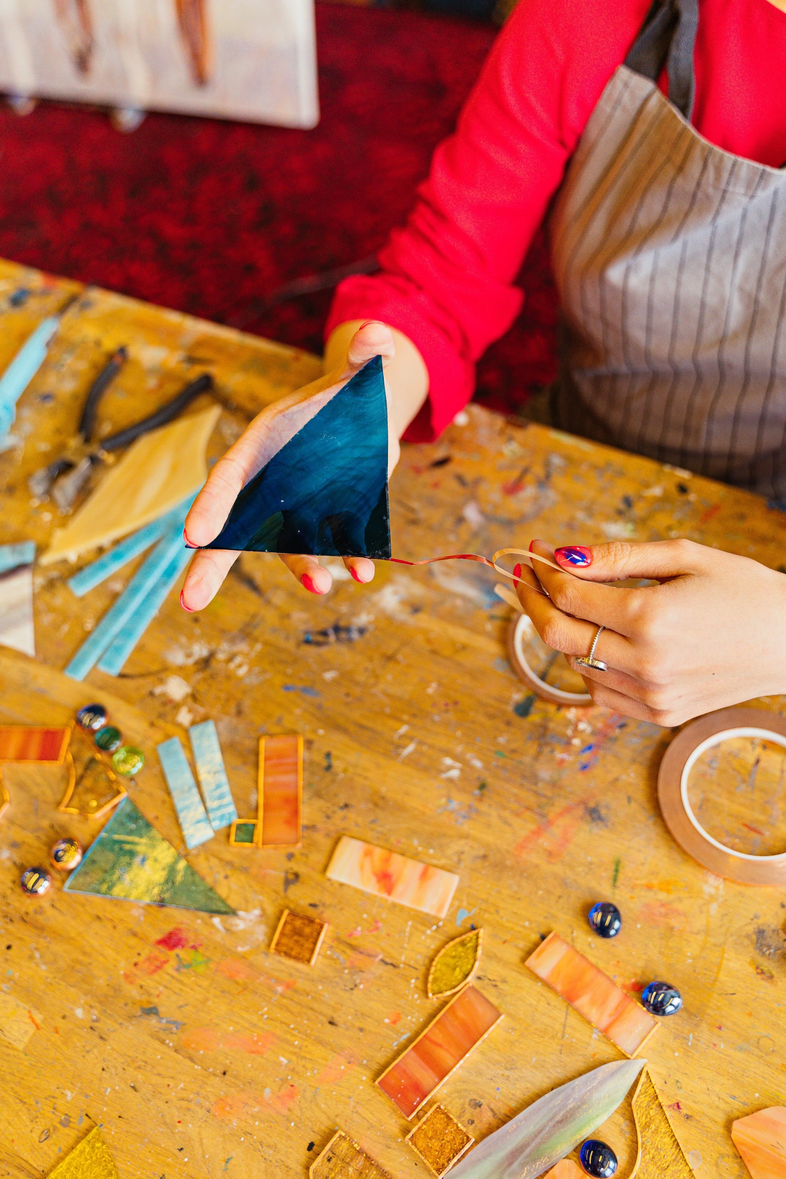 A person in a quilted apron holds a triangular stained glass piece in front of them. The workspace has various colorful glass pieces, tools, and materials on a wooden table.
