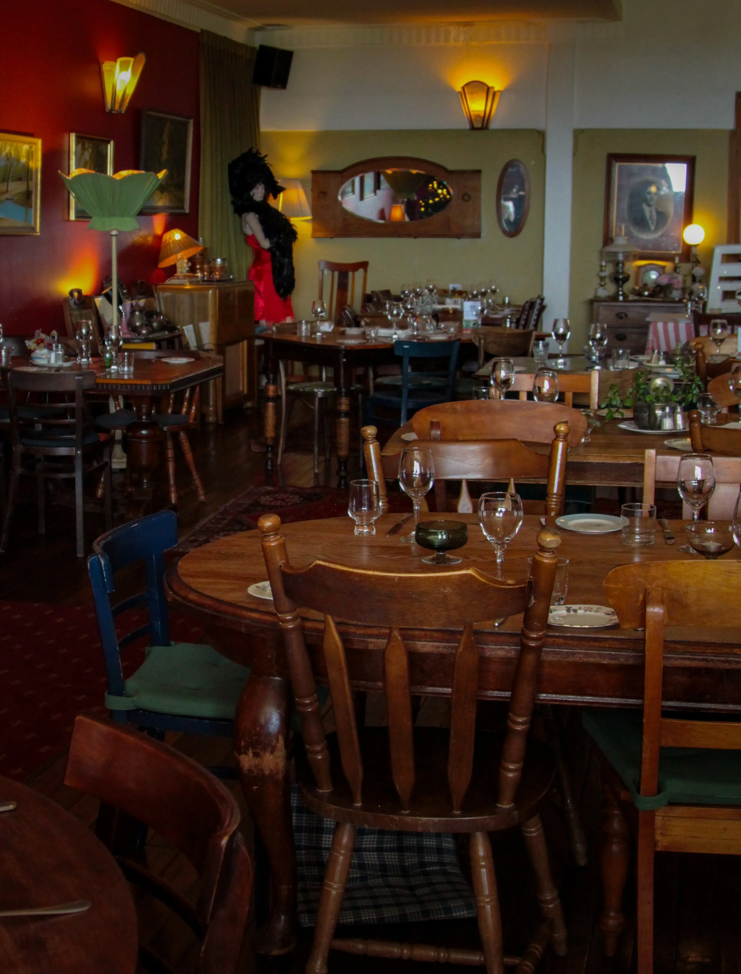 An empty restaurant dining area with several wooden tables and chairs, some set with glassware and plates. In the background, a person dressed in a black feathered costume with a red dress and black fur stole stands near the wall. The restaurant has 
