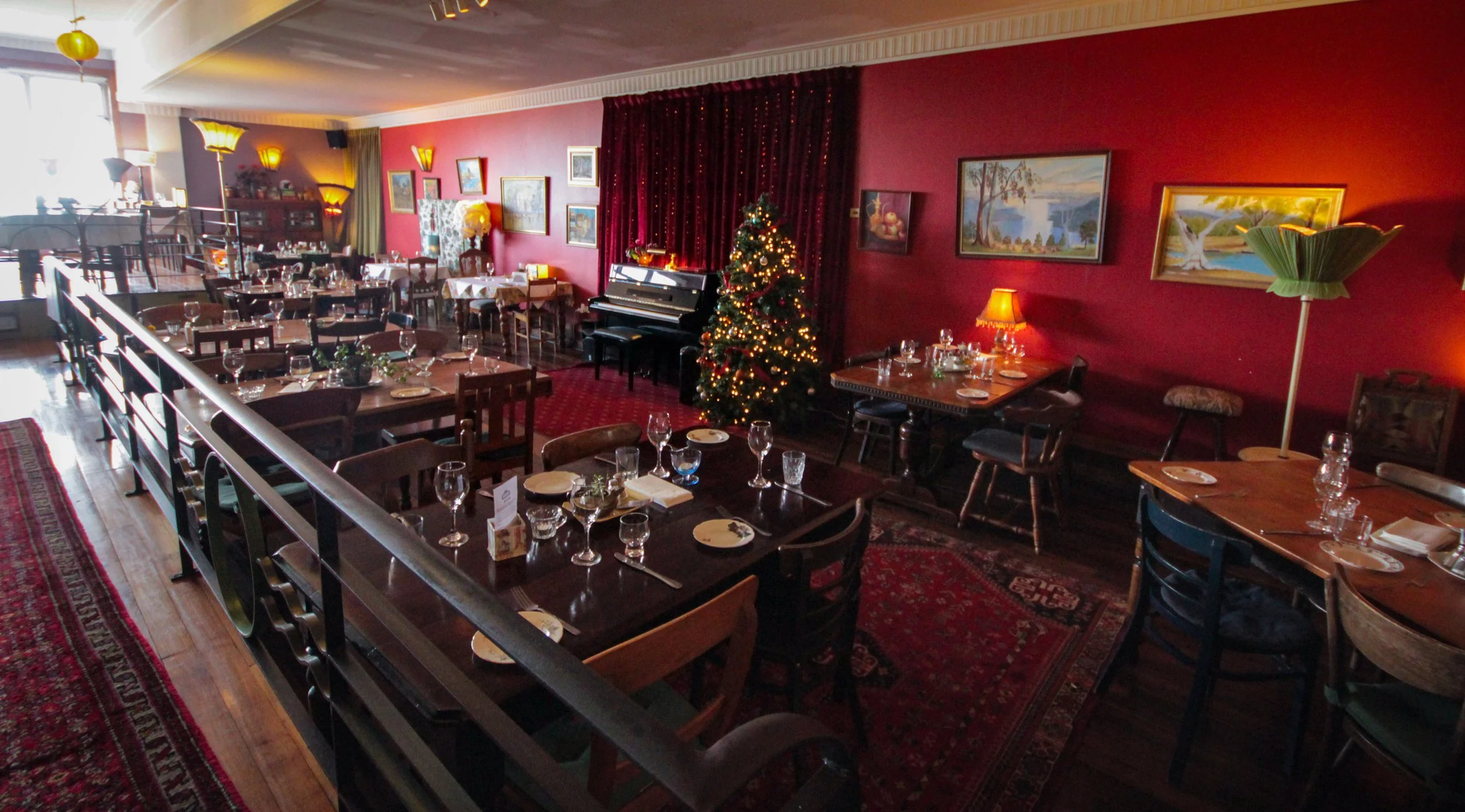 A cozy restaurant decorated for Christmas with a lit Christmas tree, paintings on the red walls, and tables set for a meal.