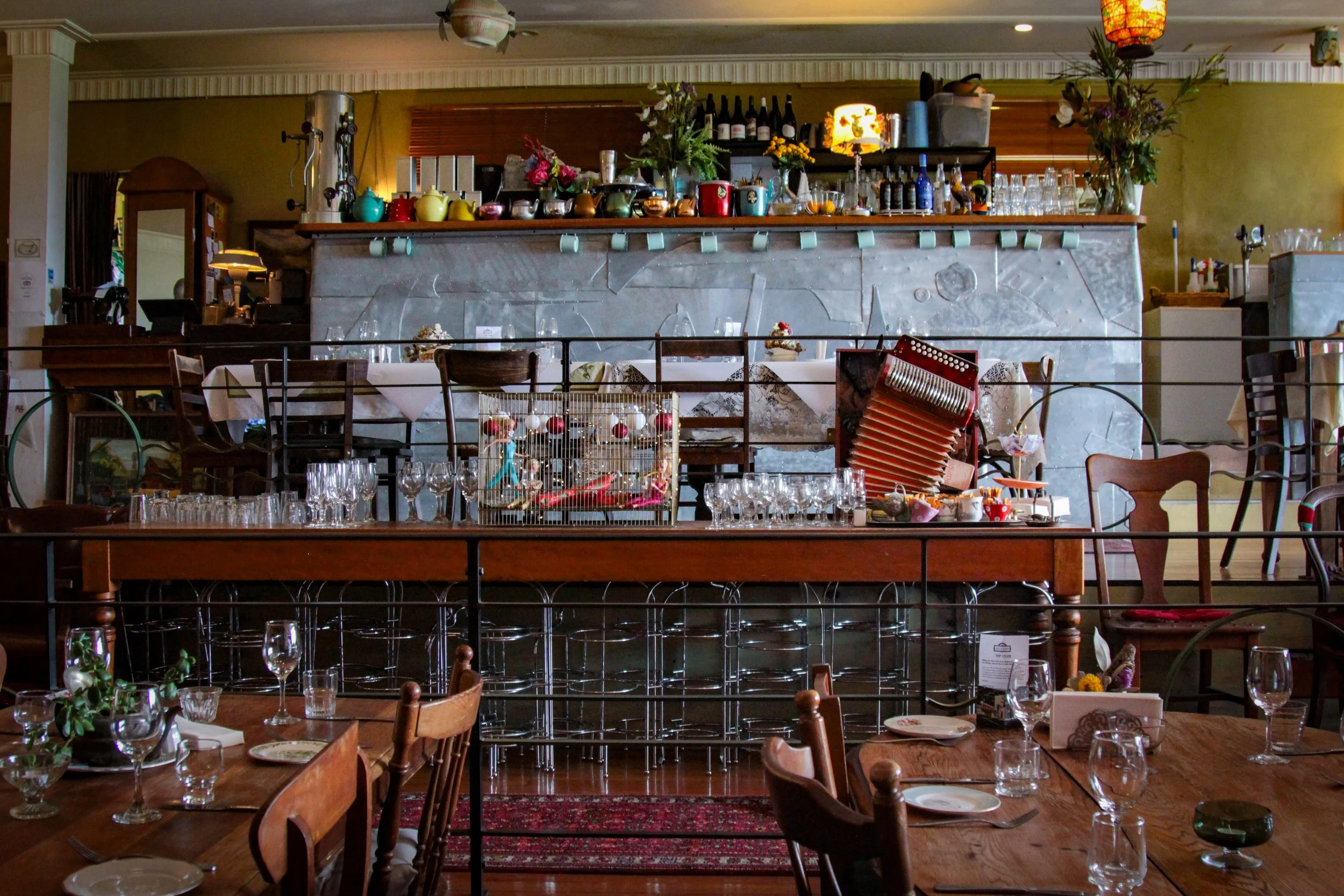 An empty restaurant with tables set for dining, a front bar area with glassware, and colorful kitchenware on shelves, decorated with flowers and lanterns.