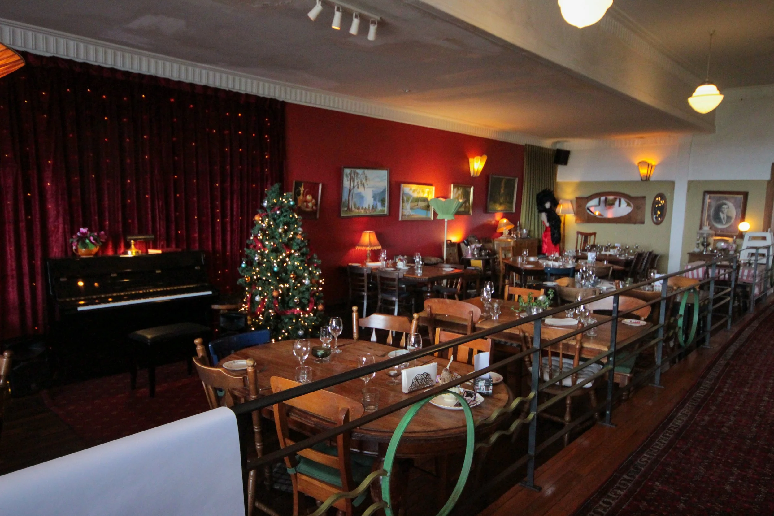 A dining area decorated for Christmas with a Christmas tree, paintings on the red wall, and tables set with glasses and plates.