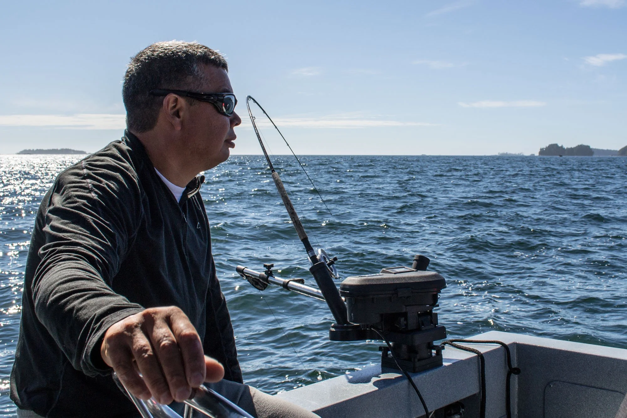 A man wearing sunglasses and a dark jacket is fishing from a boat in the ocean on a bright day. He is holding a fishing rod, and the horizon shows distant landforms with a few boats visible.