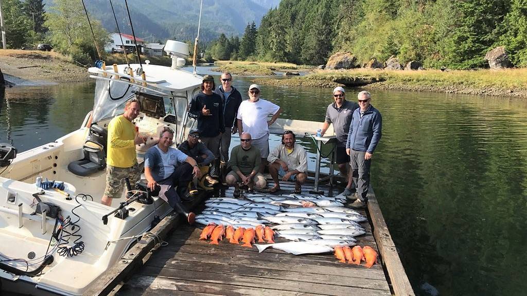 Group of people on a dock next to a boat, displaying a large catch of fish laid out on the dock, including salmon and red fish, with trees and mountains in the background.