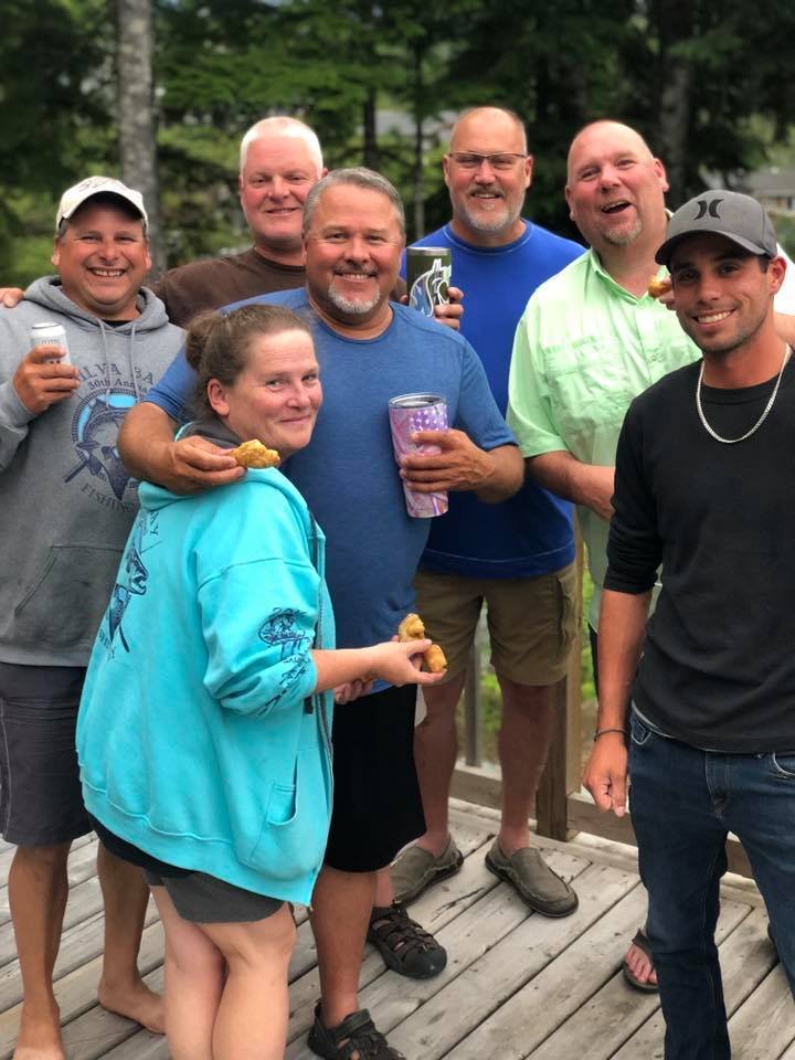 Group of seven people smiling and enjoying a casual outdoor gathering, some holding drinks and food, standing on a wooden deck with trees in the background.